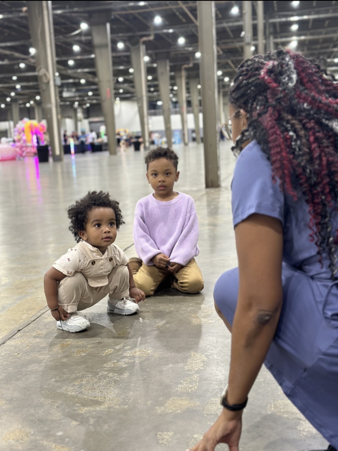 Two children sitting on the floor and a woman in scrubs kneeling in an indoor event space with high ceilings and bright lighting.