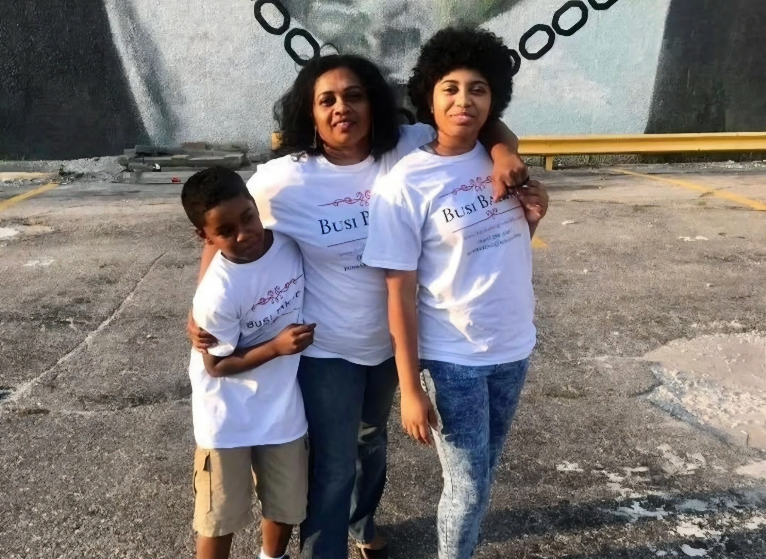 Three people standing outdoors, two women and a child, wearing matching white T-shirts, with a mural in the background.