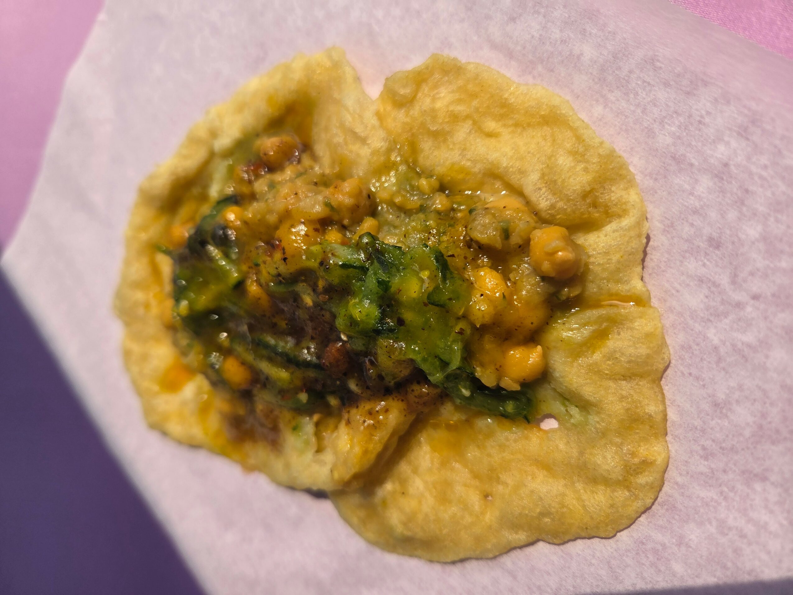 A fried snack topped with chopped green chilies and chickpeas on a pink surface.