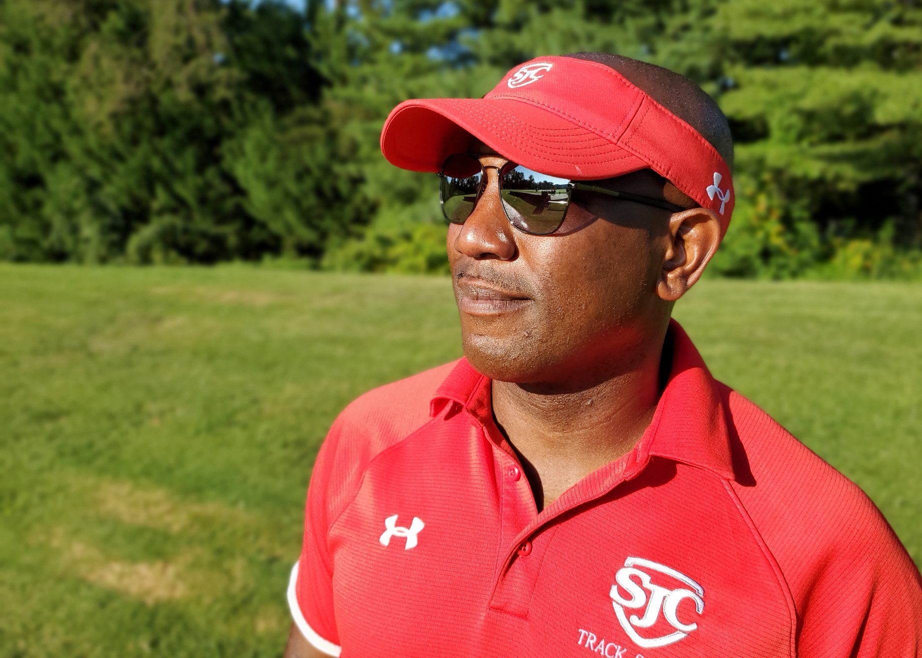 Man wearing sunglasses, red cap, and red sports shirt outdoors with green trees and grass in background.
