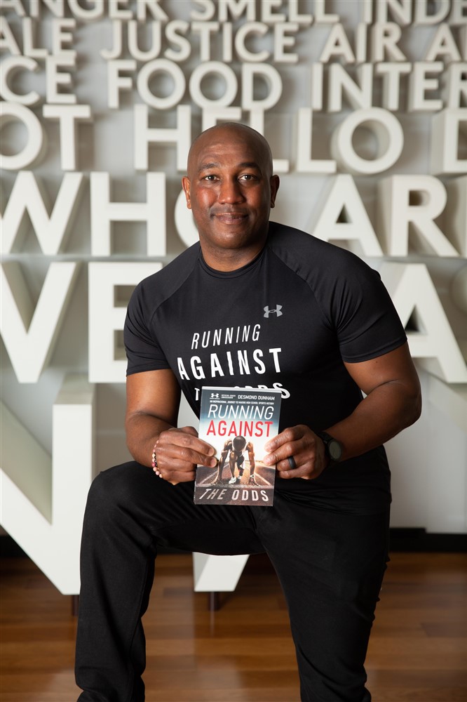 Man sitting and holding a book in front of a wall with large 3D words, wearing a black Under Armour shirt.