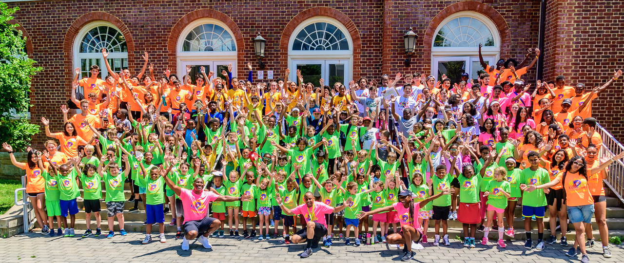 Large group of children and adults in colorful outfits posing outdoors in front of a brick building with arched windows.