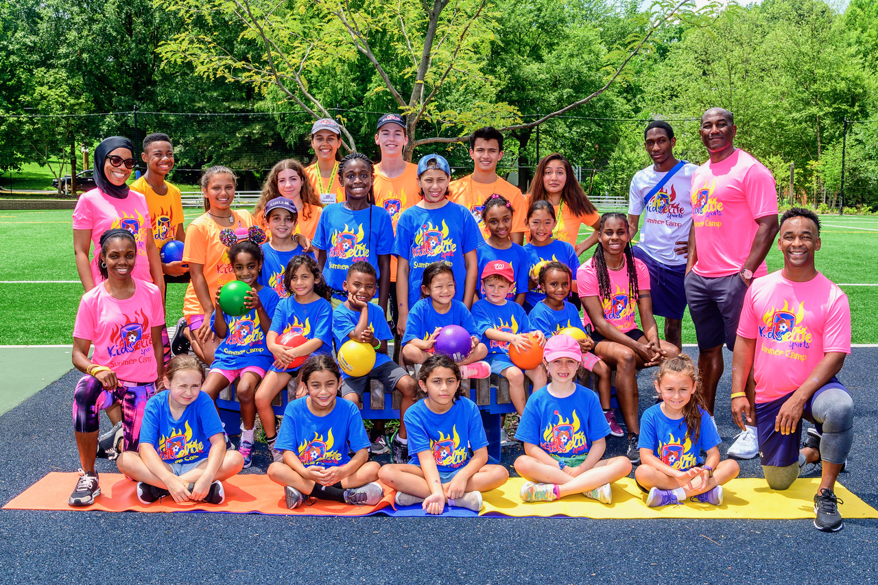 Group of children and adults outdoors on a sunny day, some holding basketballs, in colorful sportswear, on a court with trees in background.