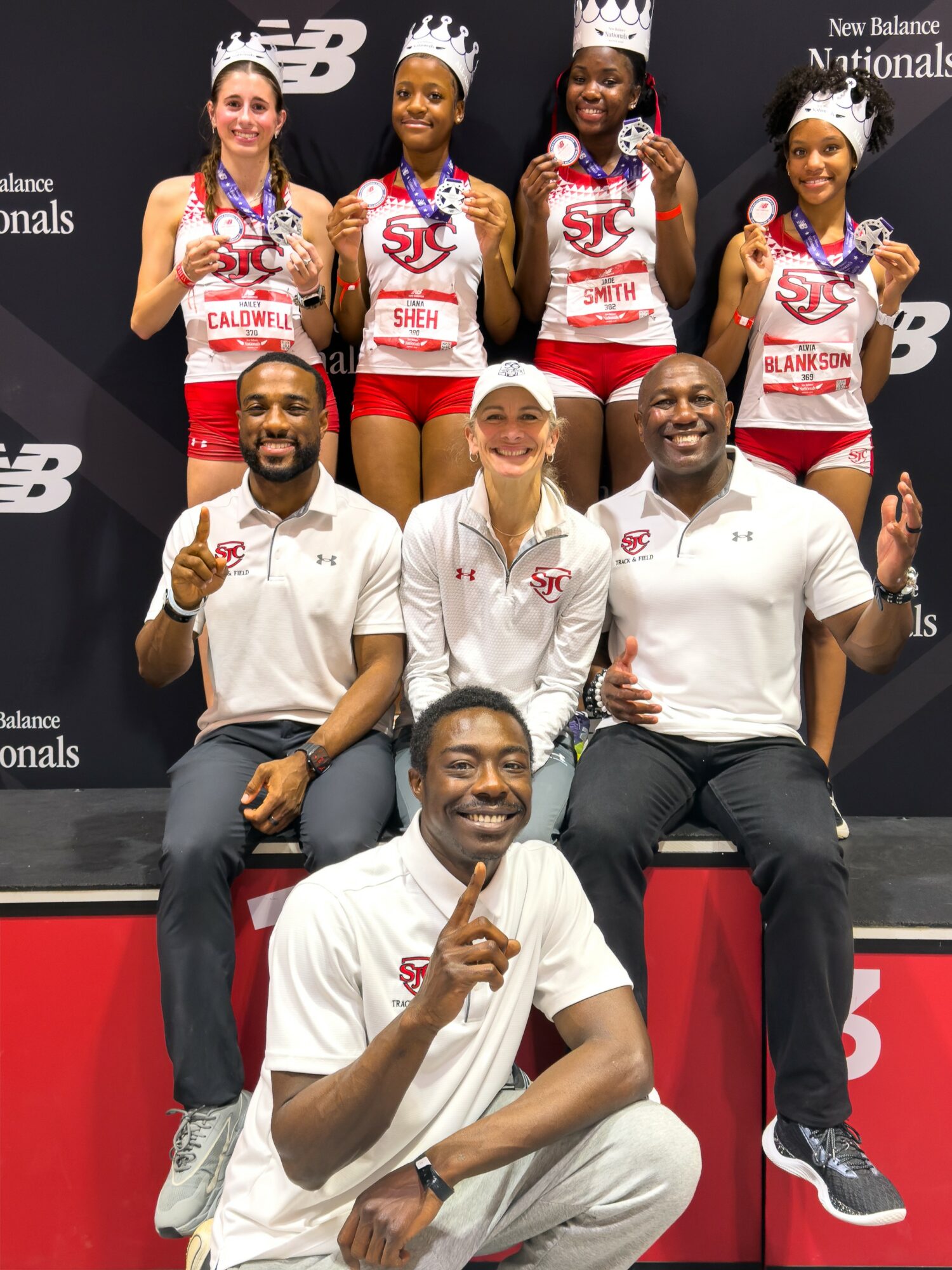 Group of female athletes in medals and crowns, with three men in front, celebrating on a podium.