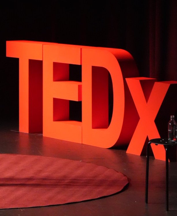 Large red TEDx sign on stage with a black background and a small table with a water bottle nearby.