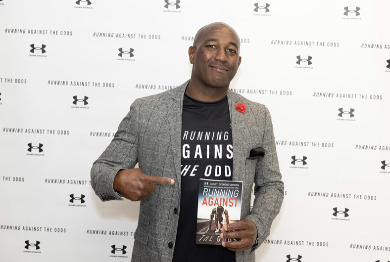 Man holding a book and pointing at it, standing in front of a backdrop with logos, wearing a gray blazer and black shirt.