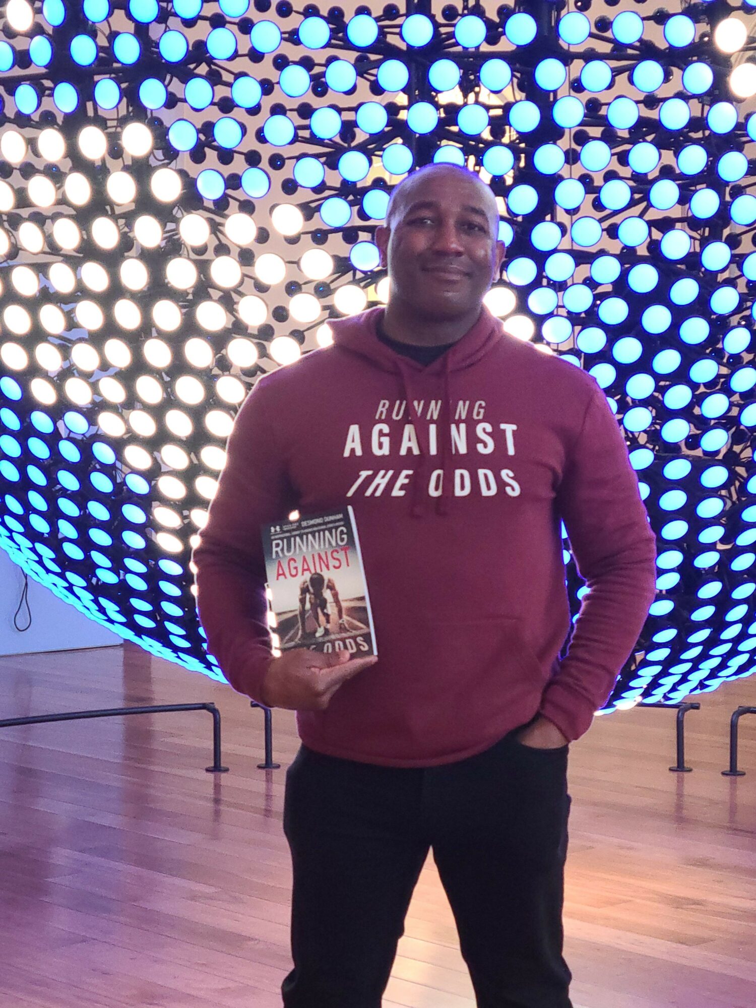 Person standing indoors in front of a circular light installation, holding a magazine, wearing a maroon sweatshirt with white text.