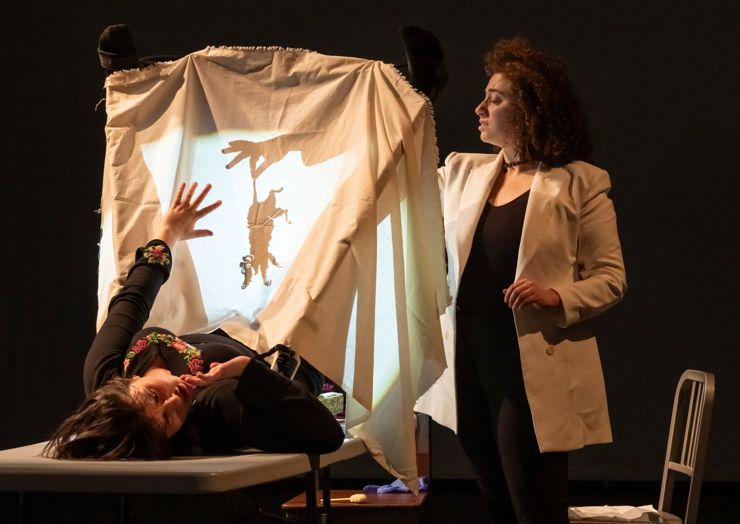 Two women on stage with a puppet show setup, one lying on a table and the other standing, with a puppet hanging from a cloth frame.