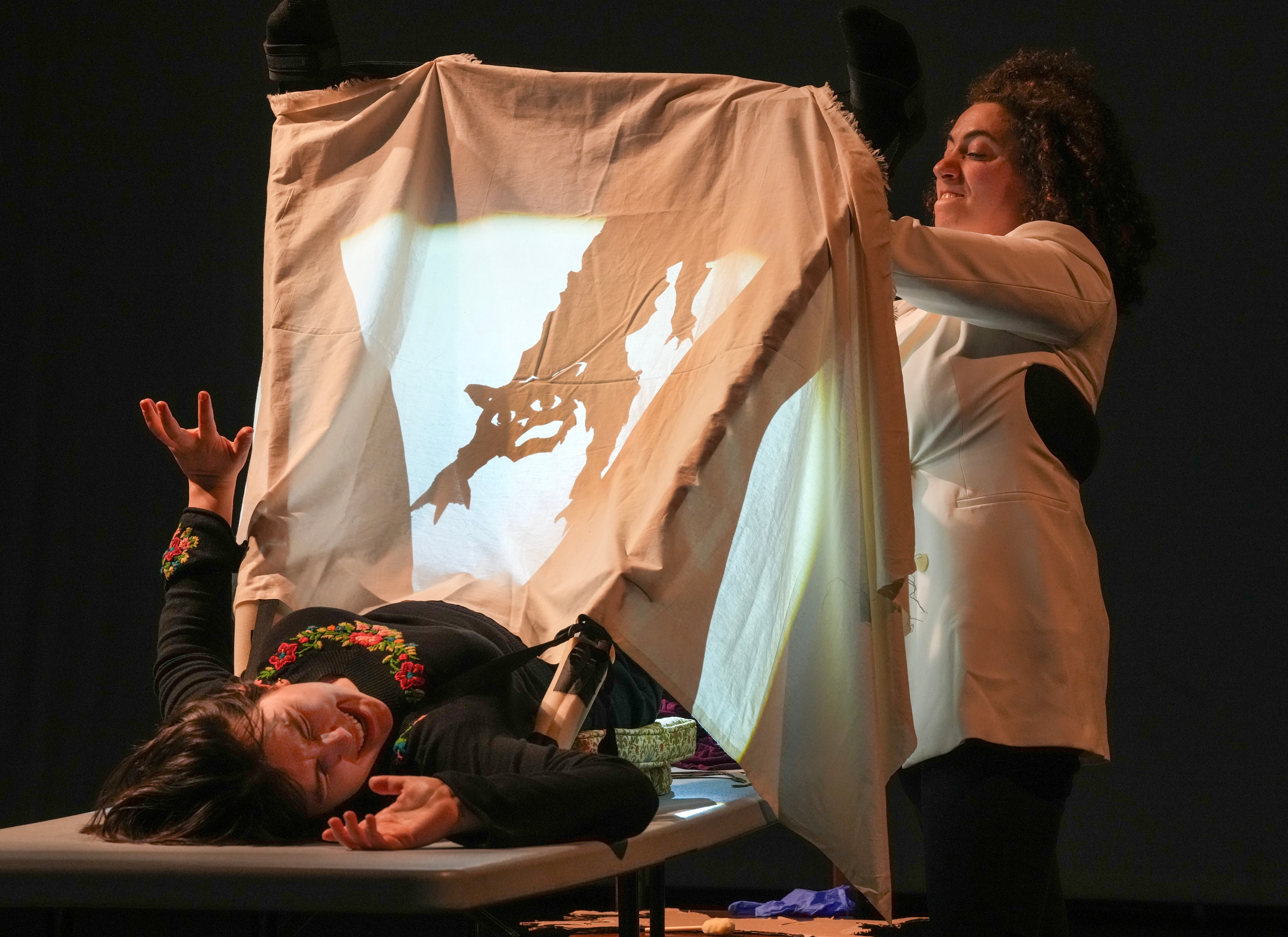 Two women perform shadow puppetry with a fabric screen, one lying down and the other standing, projecting a dragon shadow.