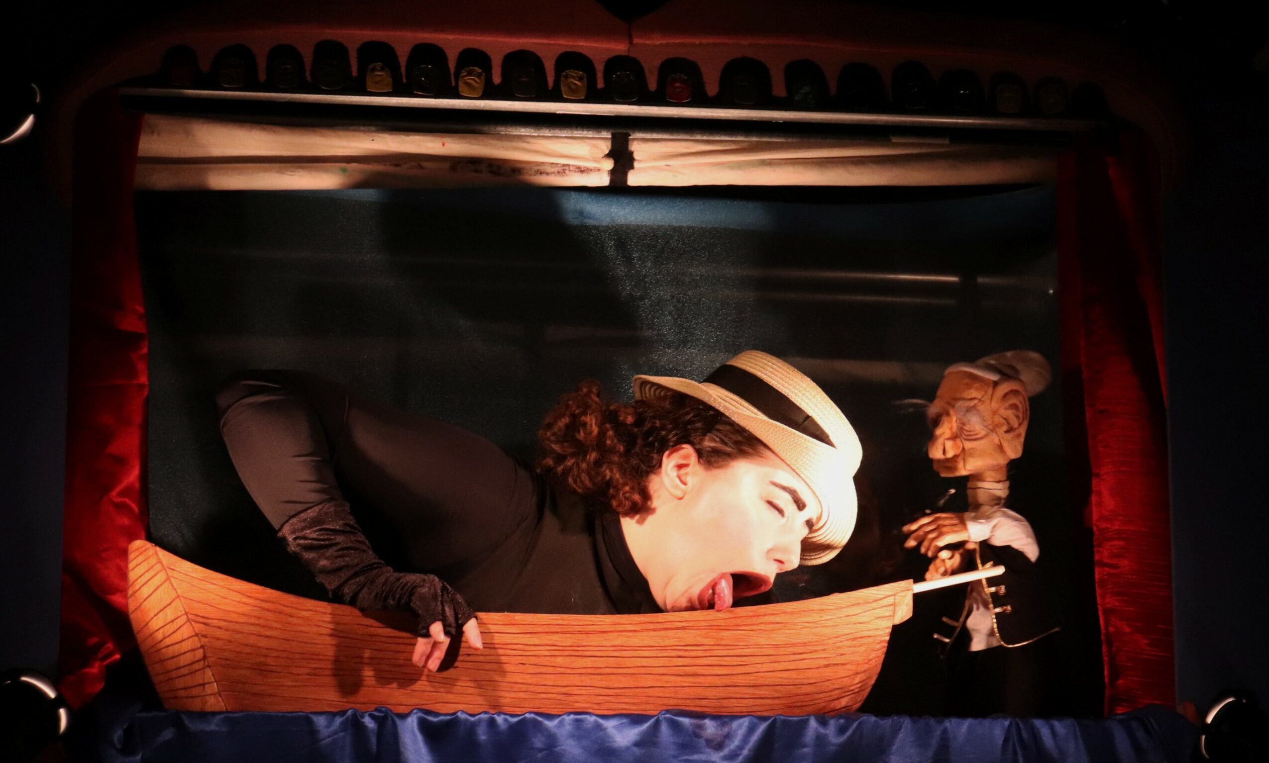 Person with curly hair and hat leaning out of a puppet theater window, holding a puppet and a glass of wine.