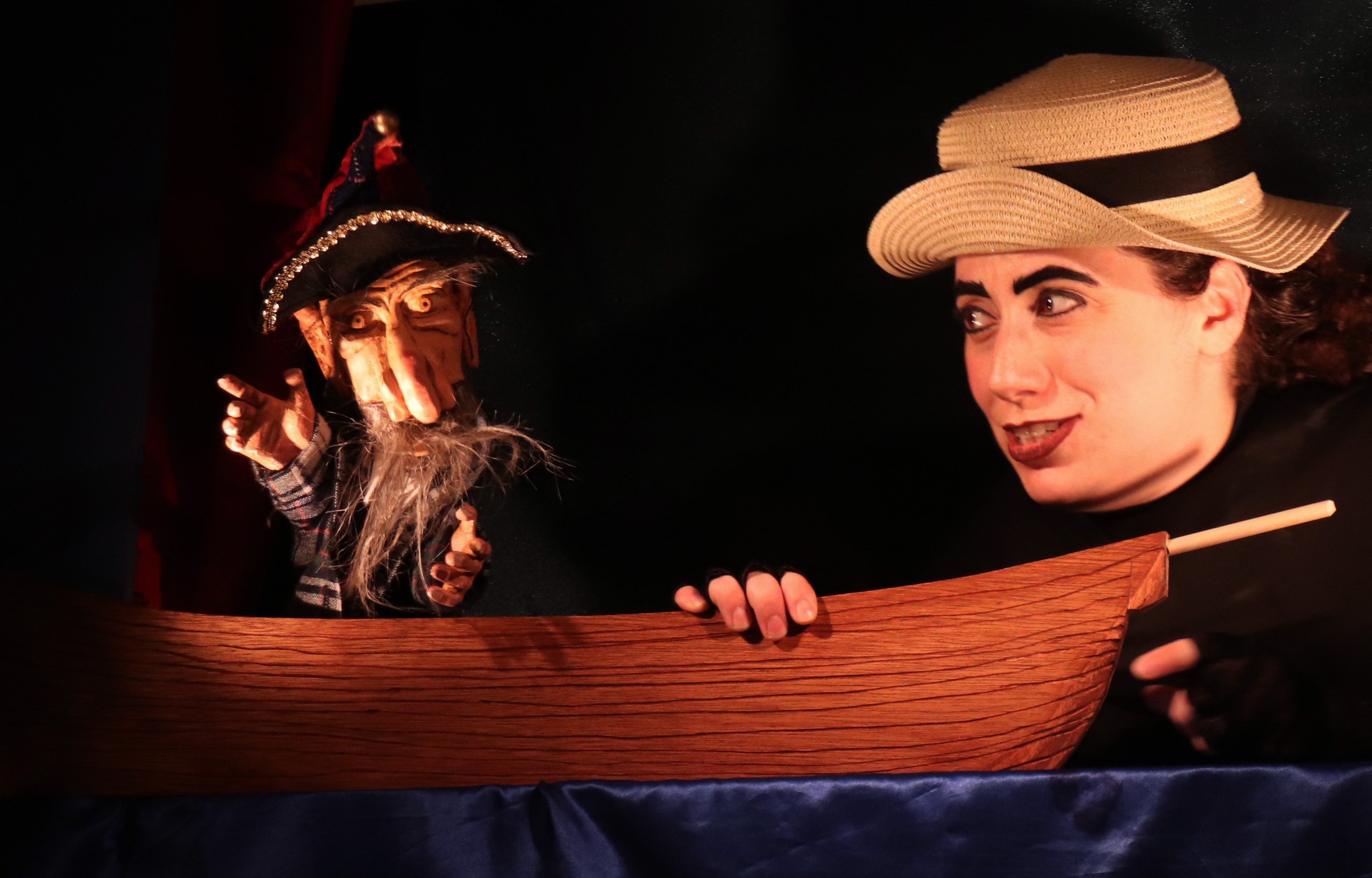 Woman with a straw hat and puppet with a hat and beard on a boat, dark background.