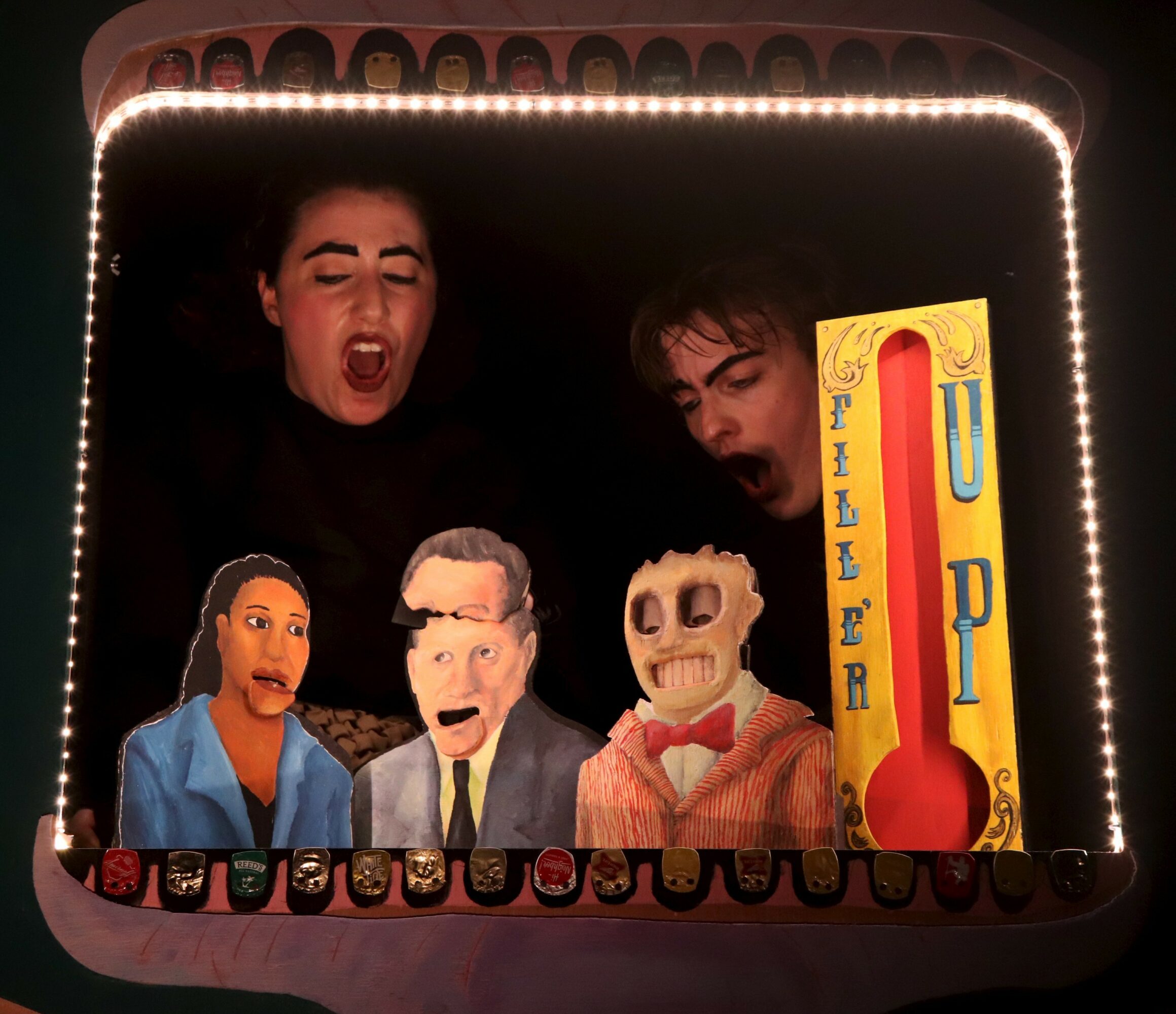Two people look at a puppet show with four puppets and a thermometer display, illuminated by bright lights.
