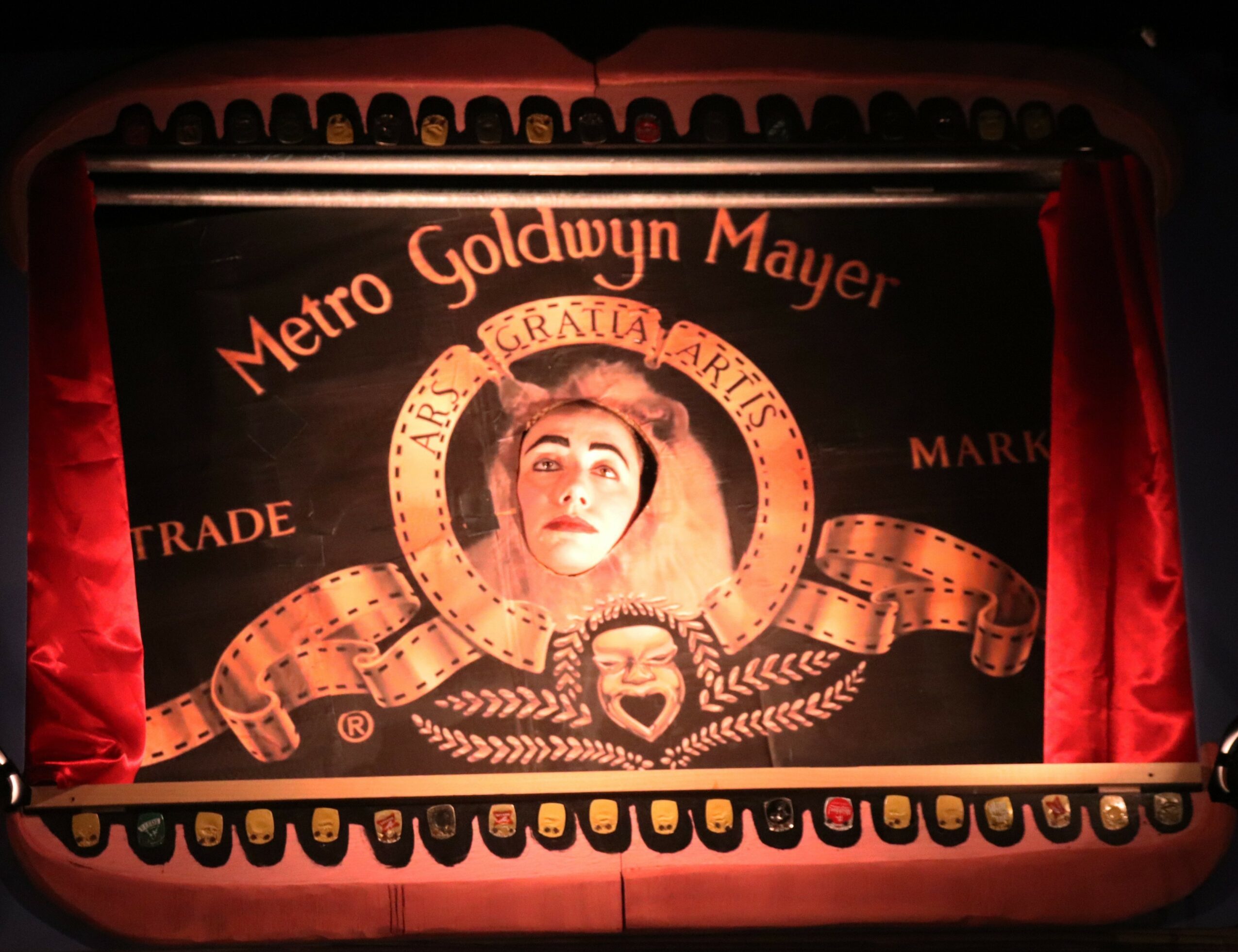 The image shows a theater stage with a curtain, a sign reading 'Metro Goldwyn Mayer,' and a lion logo with a woman's face in the center. Wordings include 'Trade' and 'Mayer.'