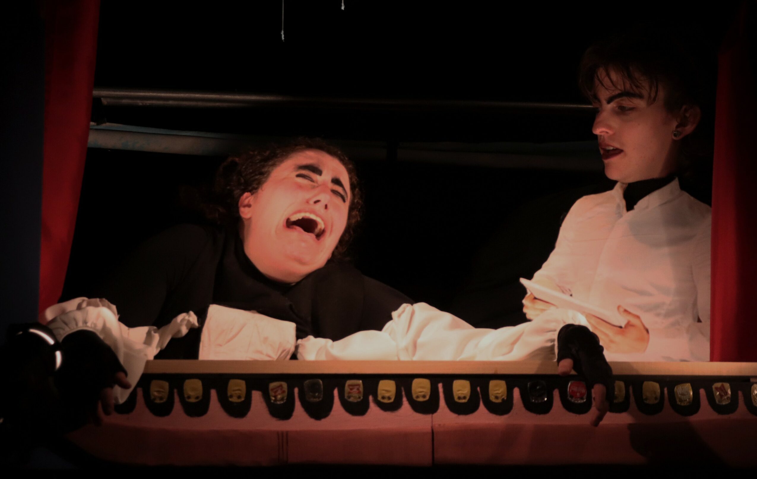 Two performers on stage, one laughing and the other looking surprised, behind a red curtain with a decorative border.