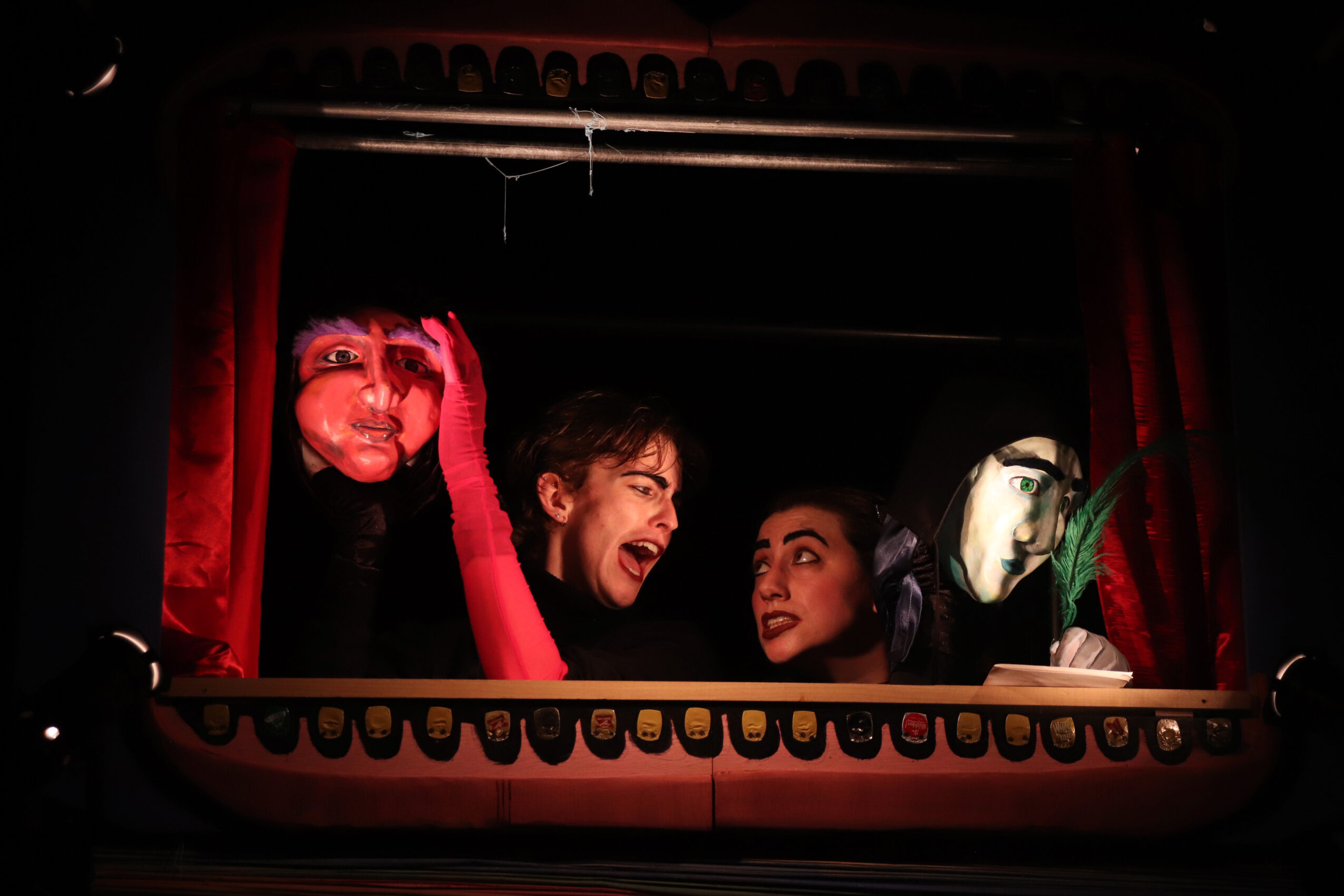 Three people look out from a theater stage with red curtains, smiling and engaging with the audience.