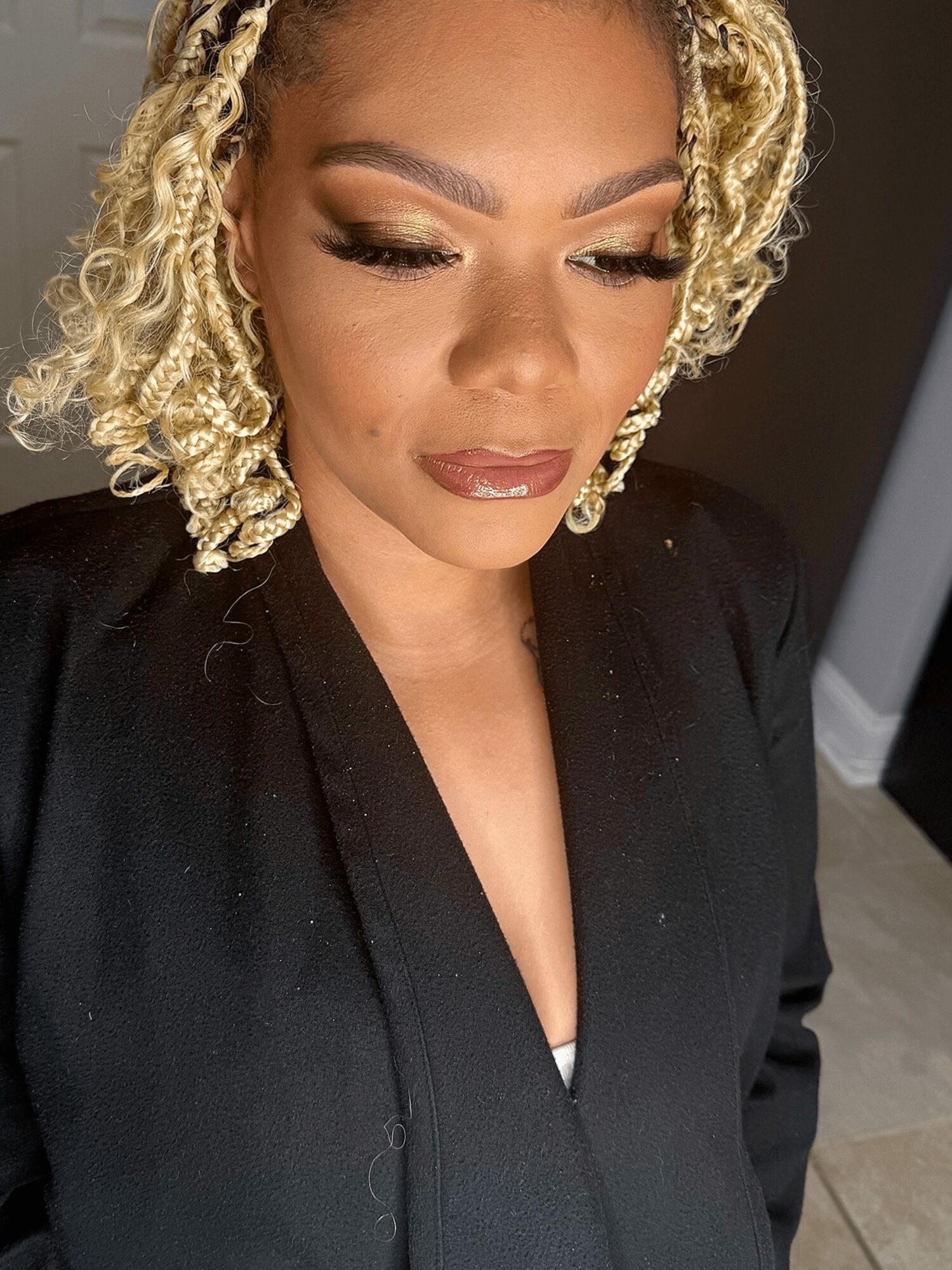 Close-up of a woman with blonde curly hair, makeup, and a black outfit, looking downward.