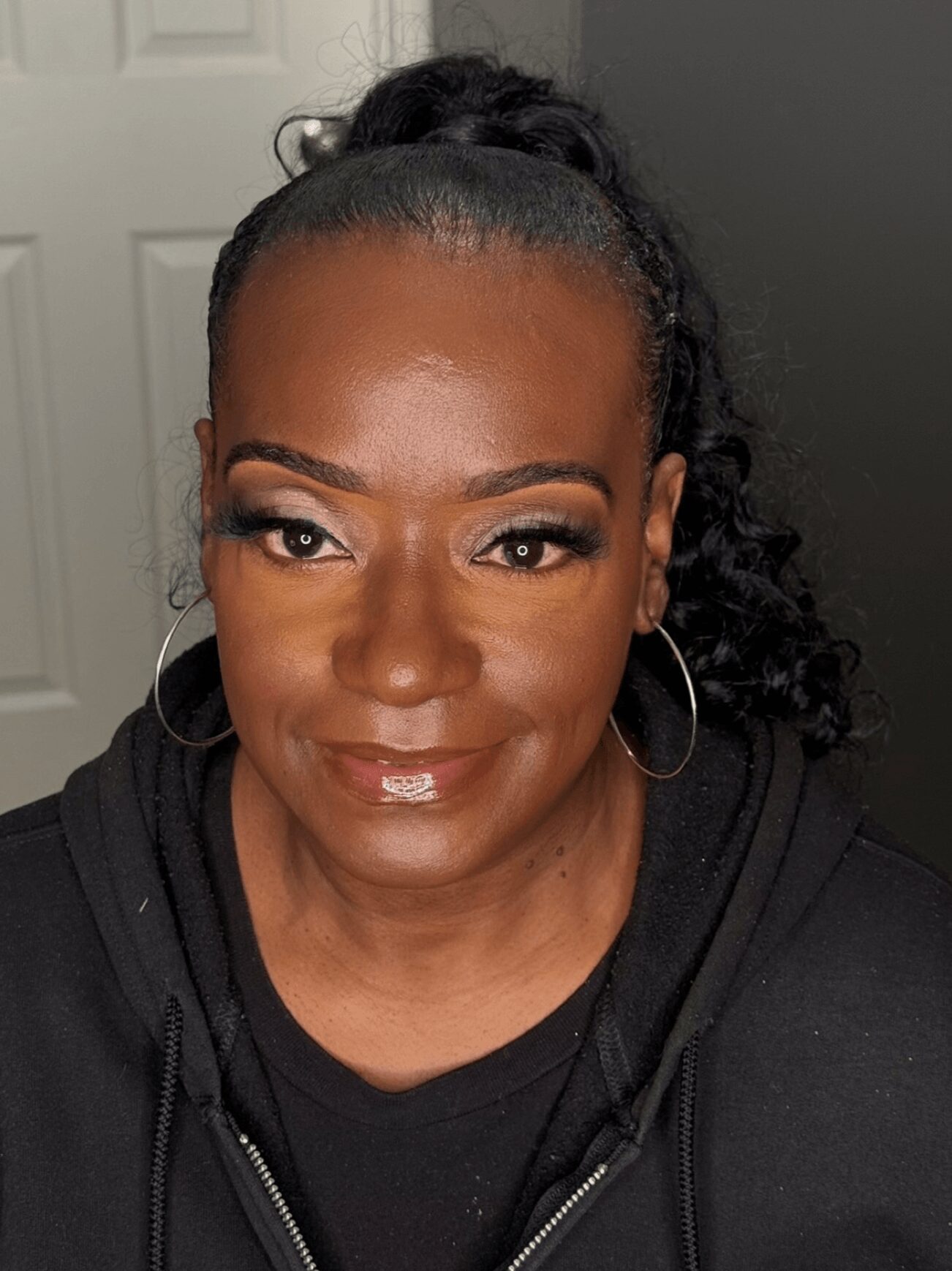 Woman with curly hair in a ponytail, wearing hoop earrings and makeup, smiling at the camera, background with a door.