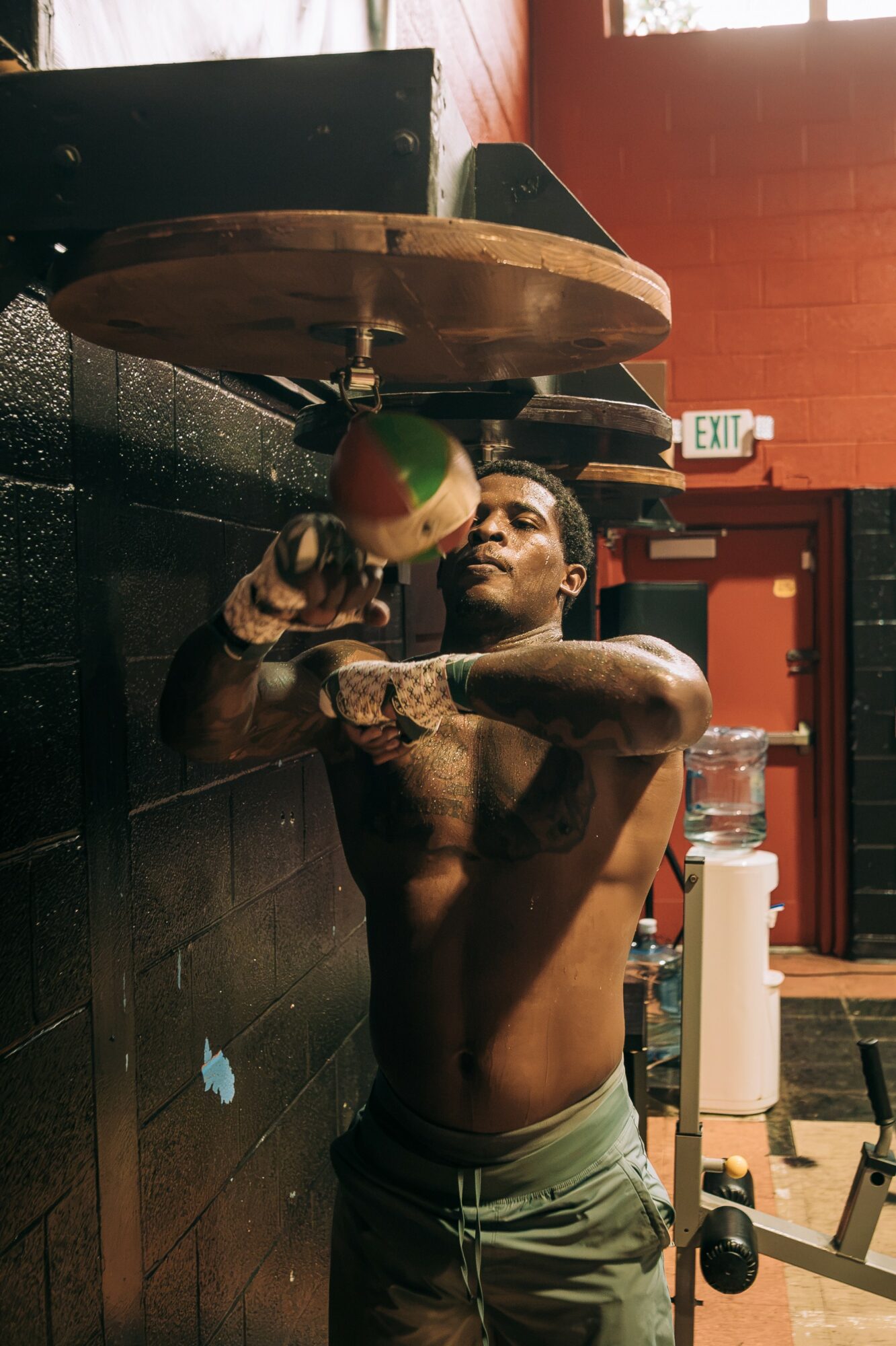 A shirtless man with tattoos throws a punch at a boxing speed bag in a gym, with a water cooler and red brick wall in background.