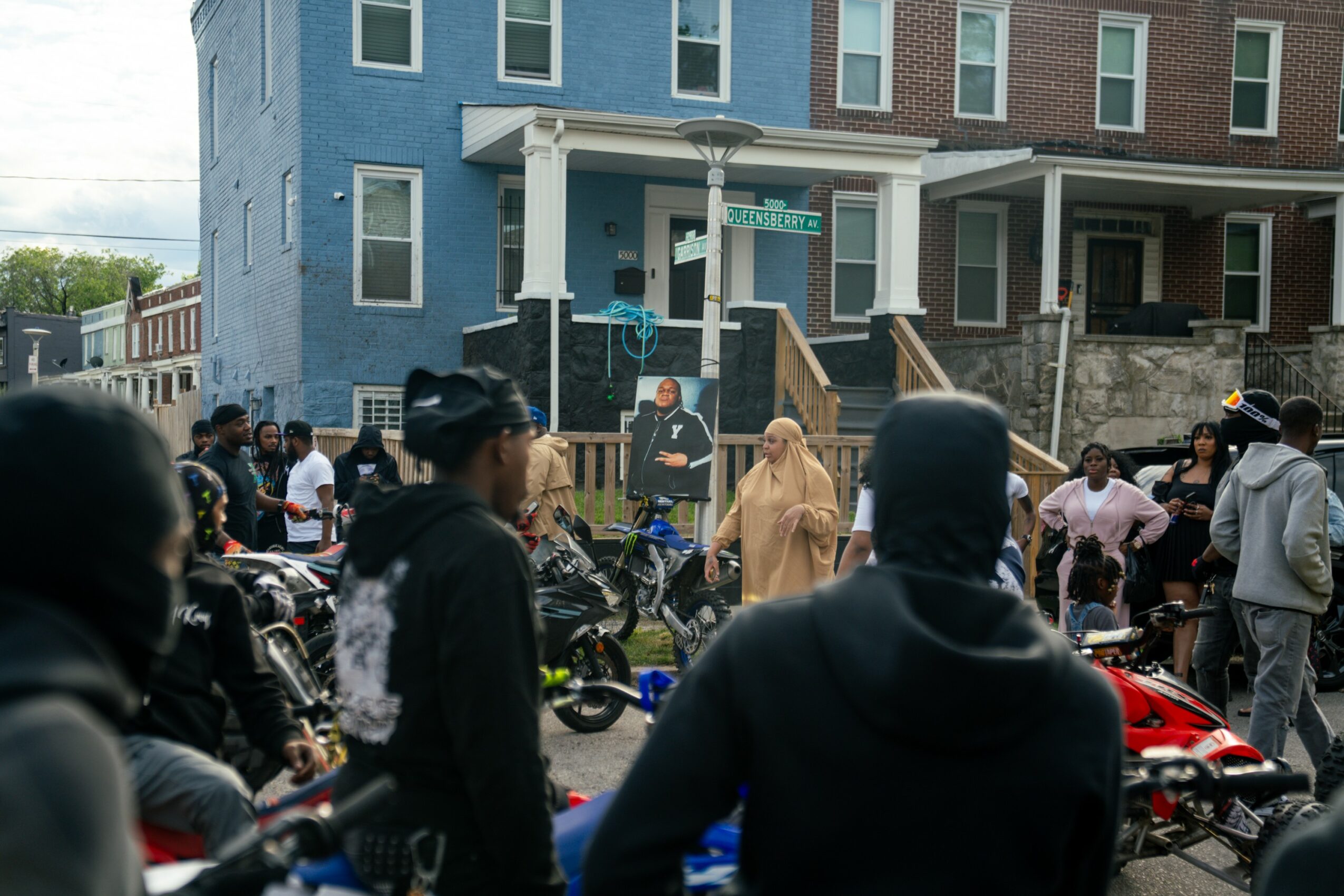 Group of people gathered outside a blue and brown multi-story building, some on motorcycles and others standing around.