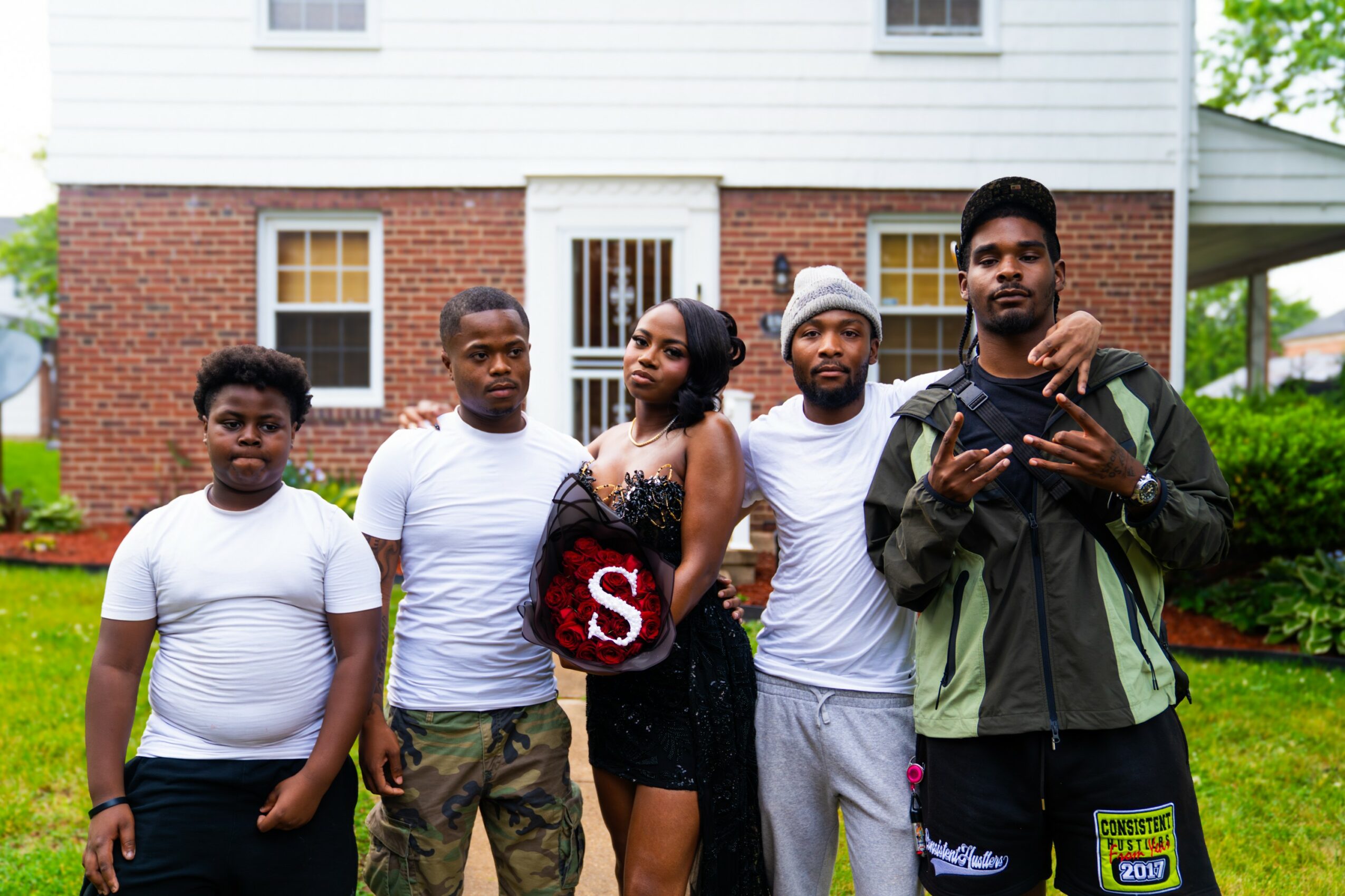 Group of five people standing outdoors in front of a house, posing for a photo, with greenery around.