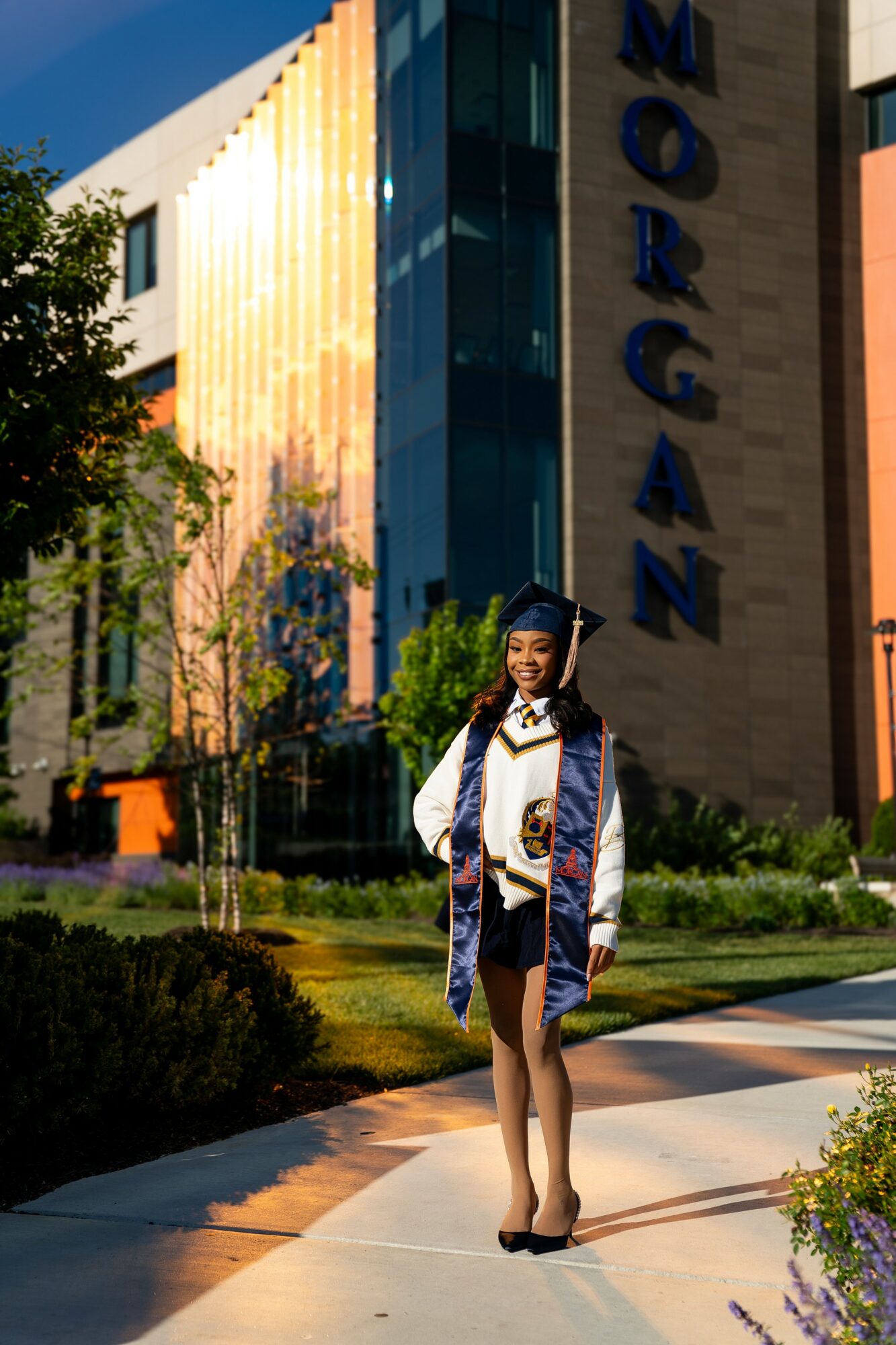 Young woman in graduation cap and gown standing outdoors near modern building with reflective glass, trees, and greenery.