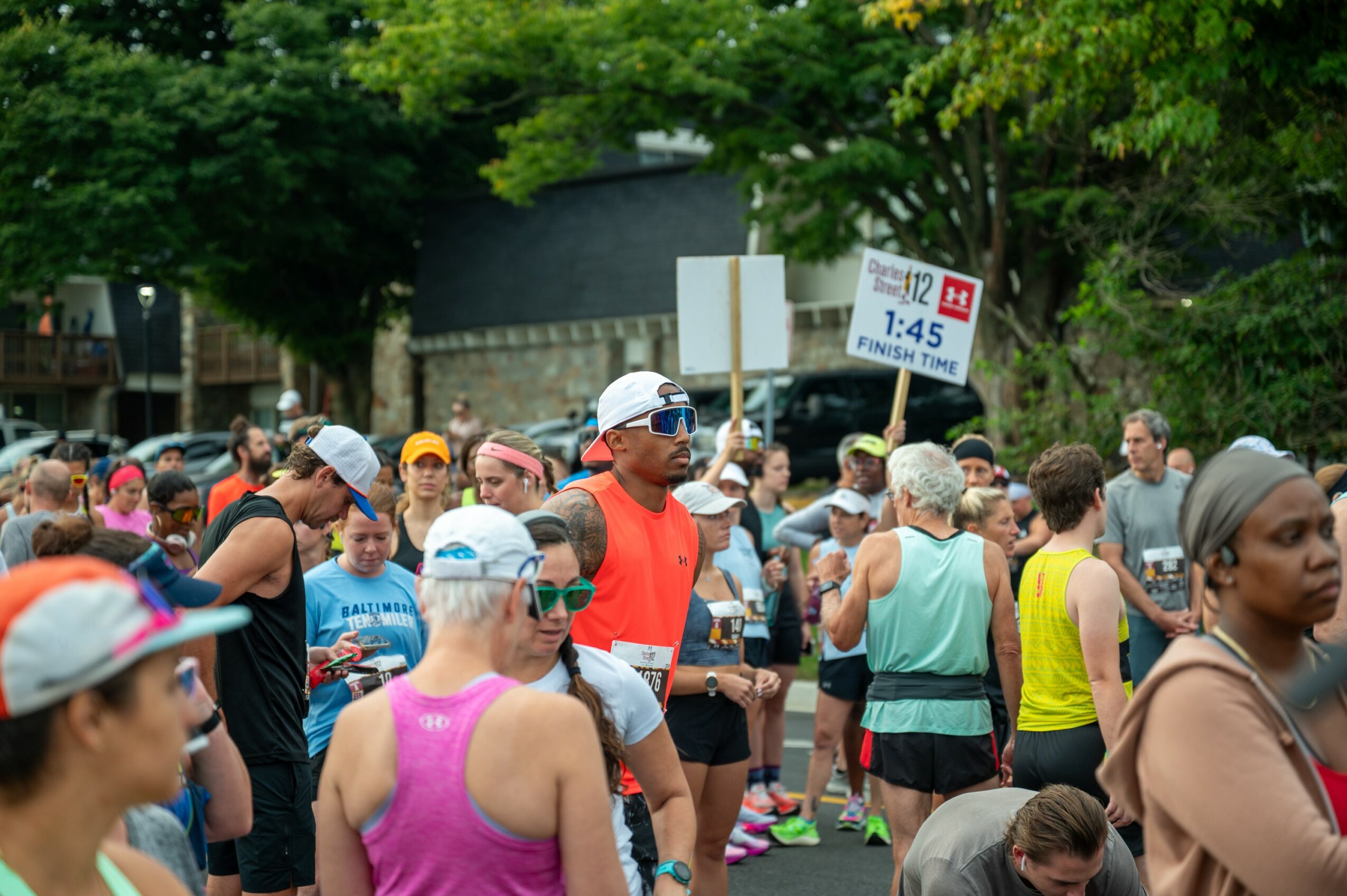 Crowd of people gathered outdoors, some wearing athletic clothing and hats, with trees and a sign in the background.
