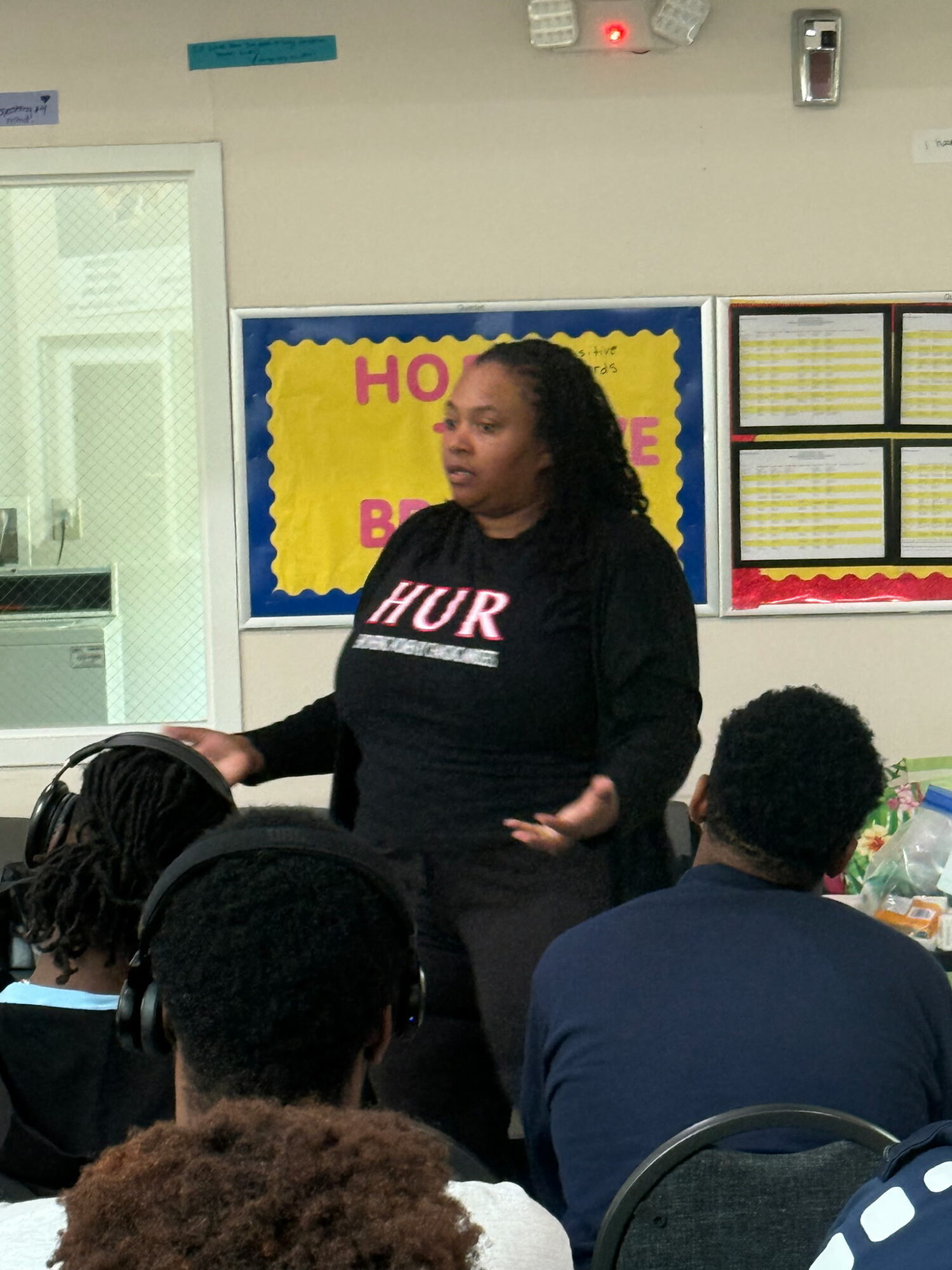 Woman speaking to a group of students in a classroom, with a yellow and blue bulletin board behind her.