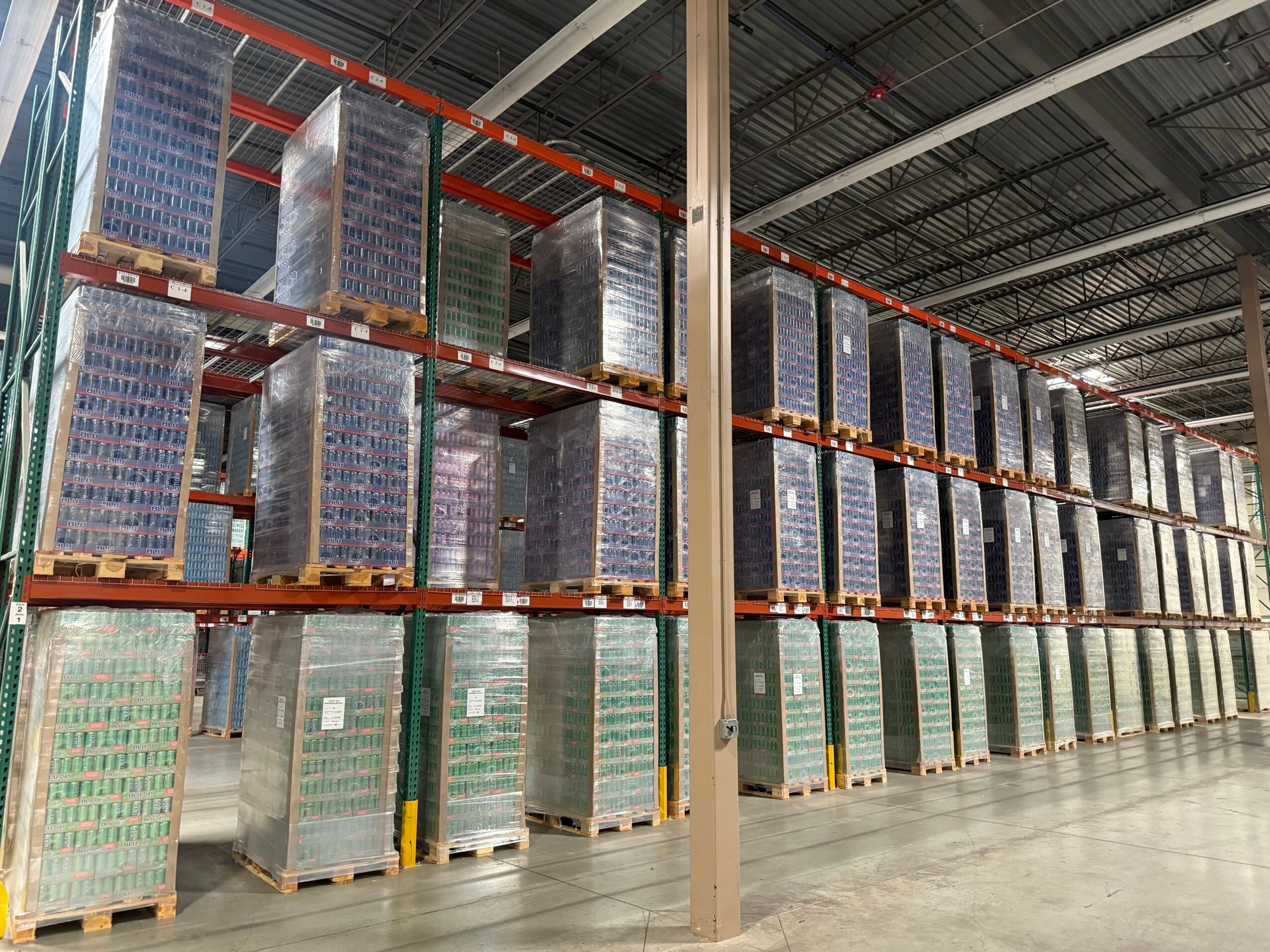 Warehouse with tall shelves stocked with boxes and pallets, high ceiling, industrial lighting, and concrete floor.