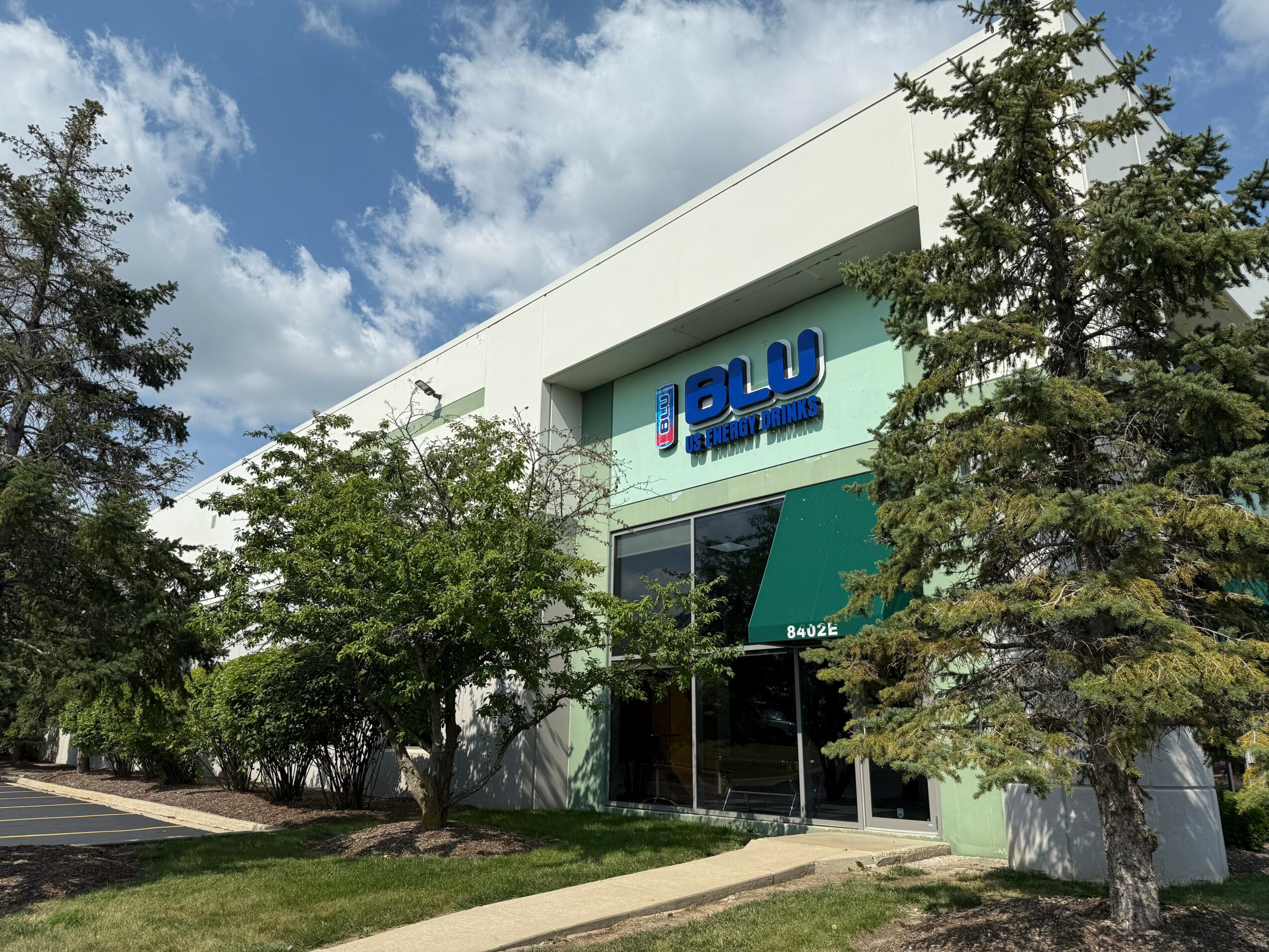 Modern building with blue and white signage, surrounded by trees and a sidewalk, under a partly cloudy sky.