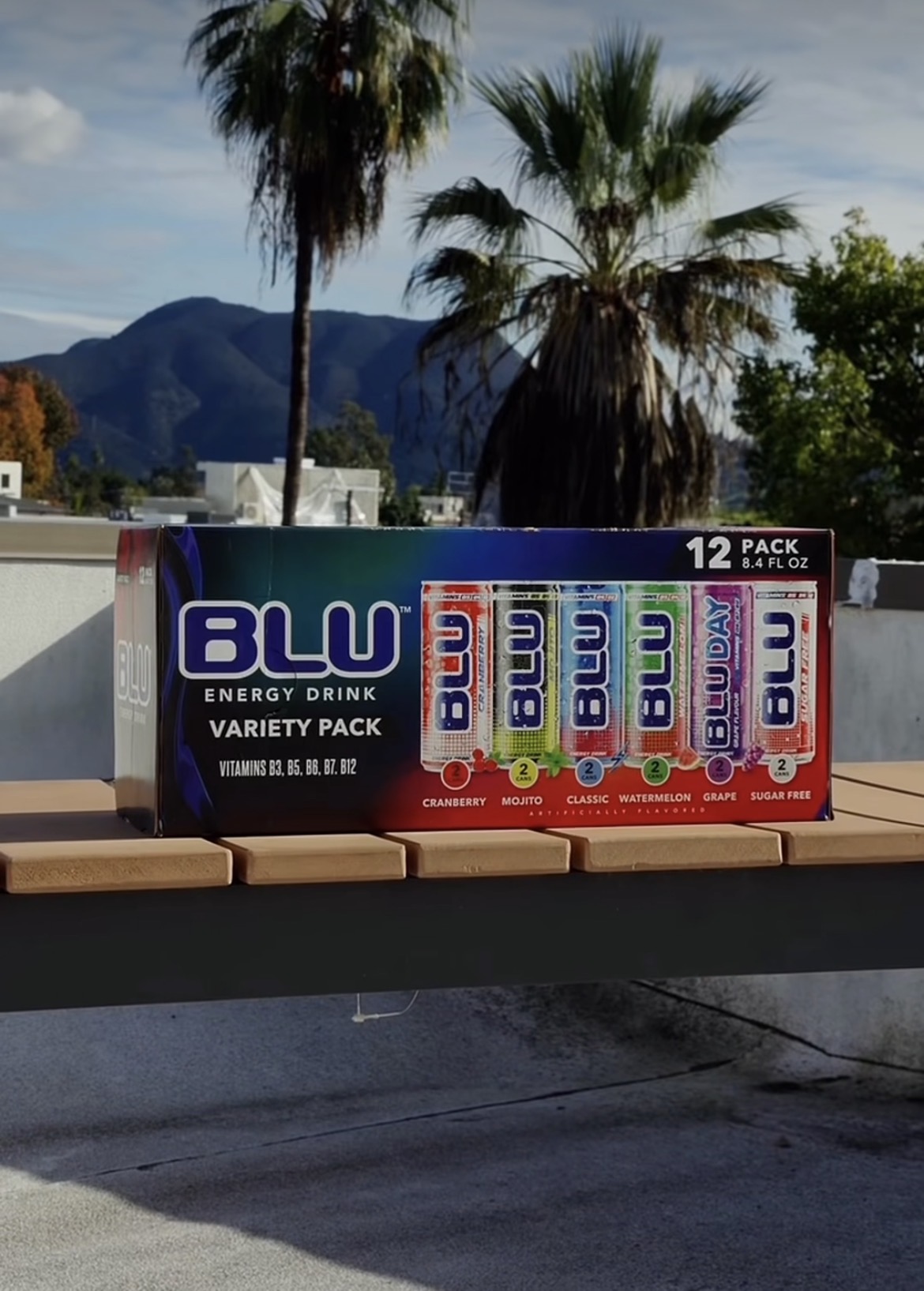 Box of 12-pack BLU energy drinks with palm trees and mountains in background, outdoors, daytime.