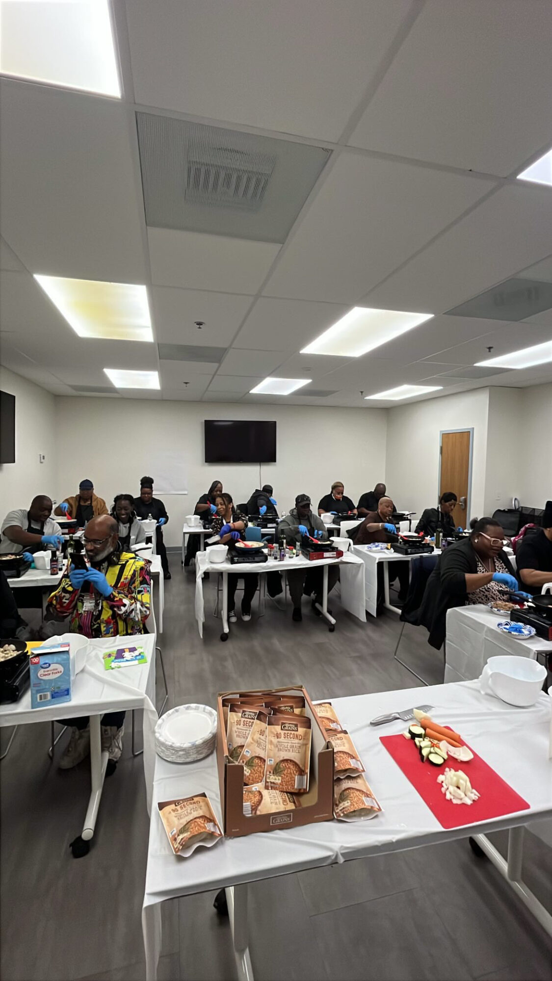 People seated at tables in a room, working or eating, with a TV on the wall, ceiling lights, and a door.