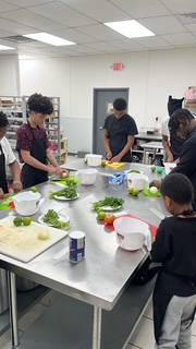 Group of children in a kitchen preparing vegetables around a stainless steel table, with shelves and kitchen equipment in background.