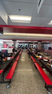 Indoor dining area with long tables and red benches, people seated, ceiling with lights, and large windows in the background.