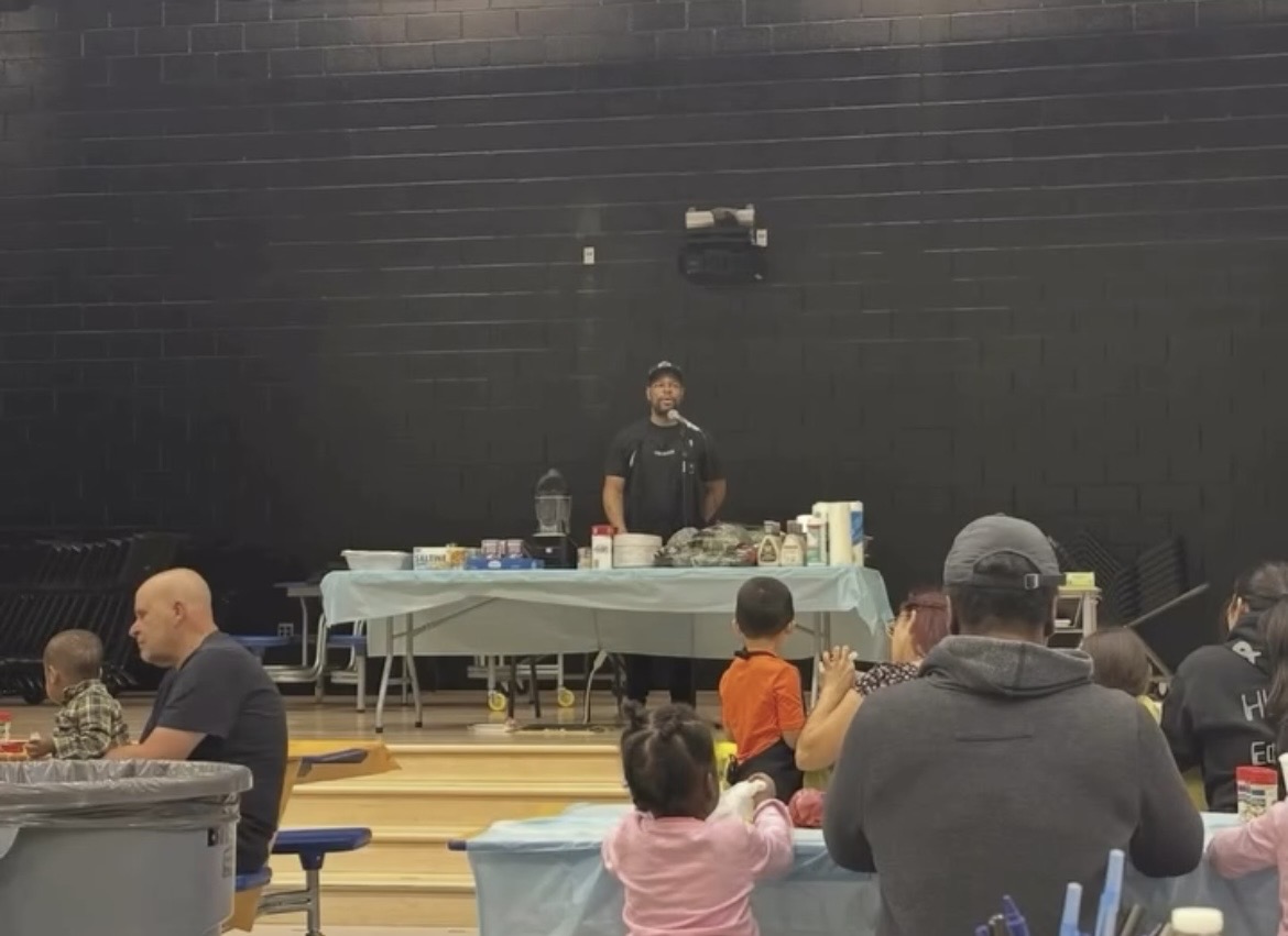 Person standing behind a table with food and drinks, people sitting at tables in a large indoor space.