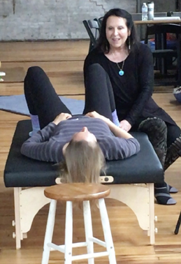 Woman sitting on a chair, woman lying on a massage table, wooden floor, indoor setting, casual clothing, smiling.