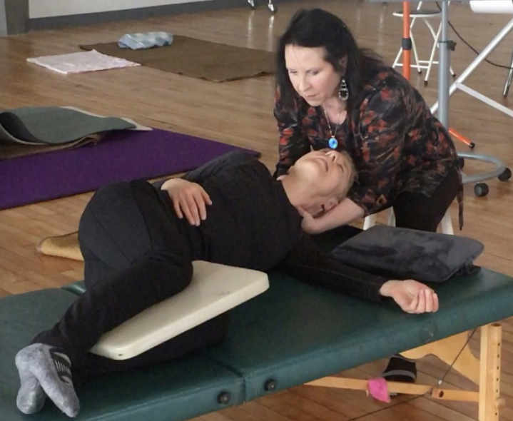 Woman supports a person lying on a therapy table during a treatment session, with therapy equipment in the background.