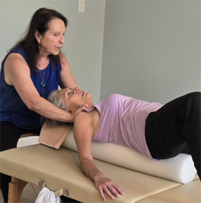 A woman provides physical therapy to an elderly woman lying on a treatment table, supporting her head and neck.