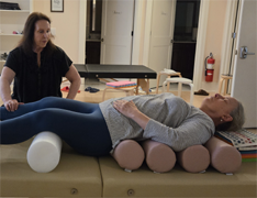 Woman lying on a treatment table with foam rollers underneath her back, in a clinical setting, with another woman nearby.