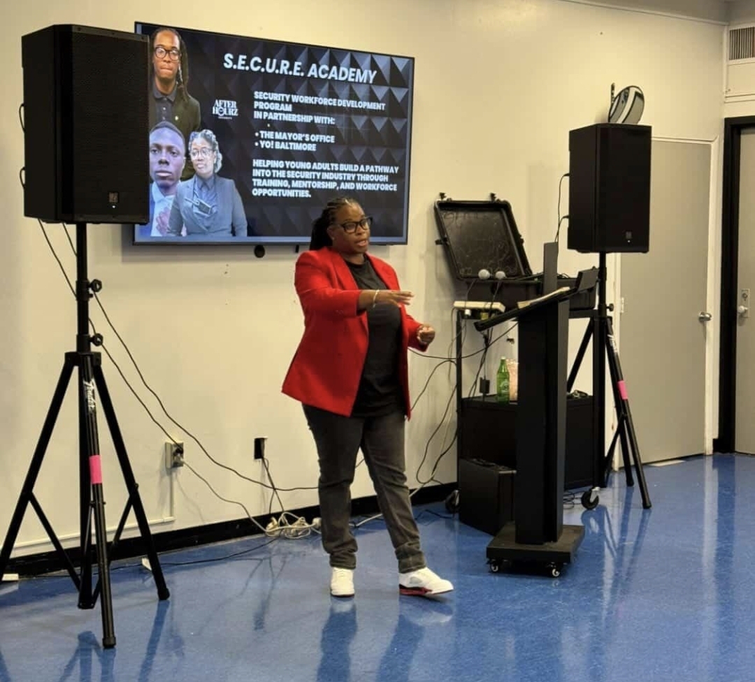 Woman in red jacket speaking in front of a screen with text and images, in a room with blue floor and speakers.