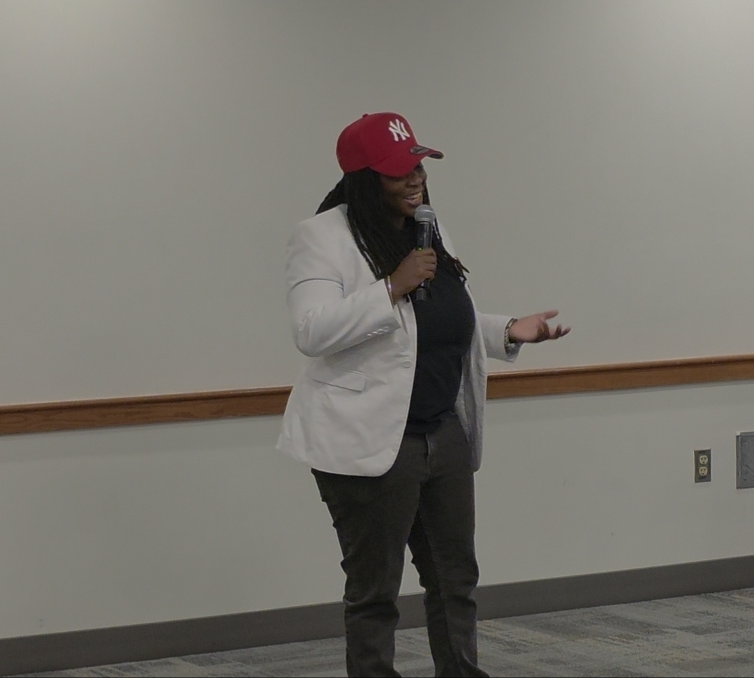 Woman speaking into microphone, wearing a red cap, white blazer, and black pants, standing in front of a plain wall.