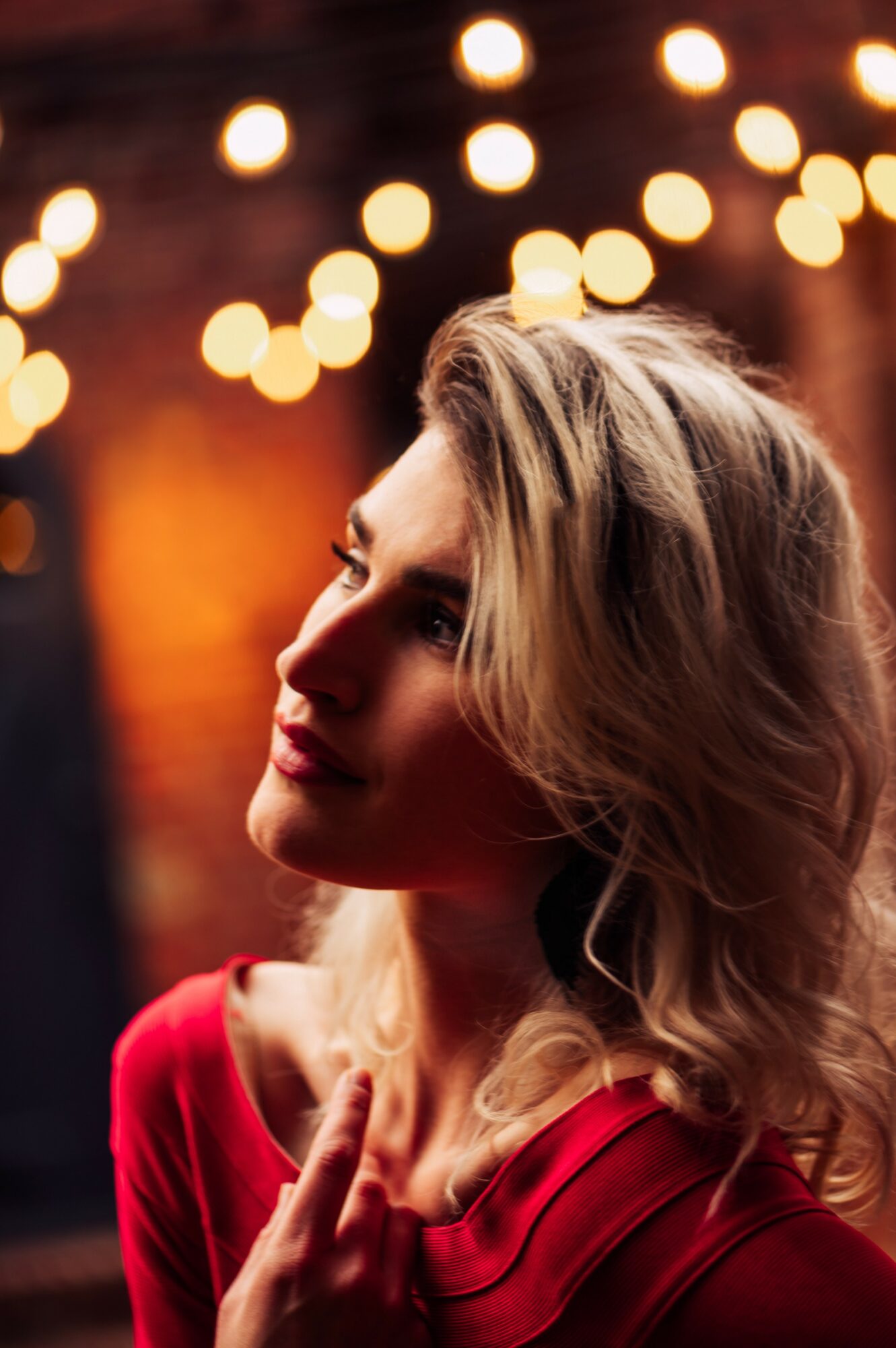 Side profile of a woman with blonde hair wearing a red top, illuminated by warm bokeh lights in the background.