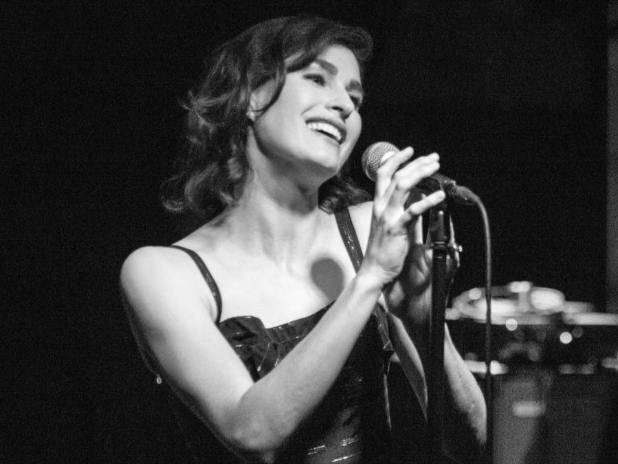 Woman singing into a microphone, smiling, with a piano in the background, in black and white.