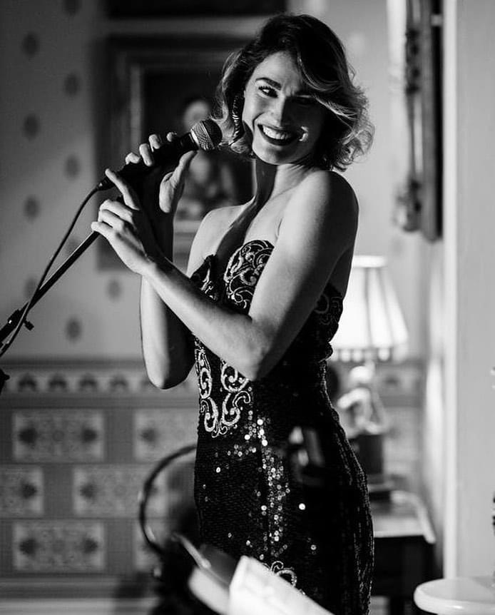 A woman with wavy hair holding a microphone, smiling, wearing a sequined dress, in a decorated room with a lamp.