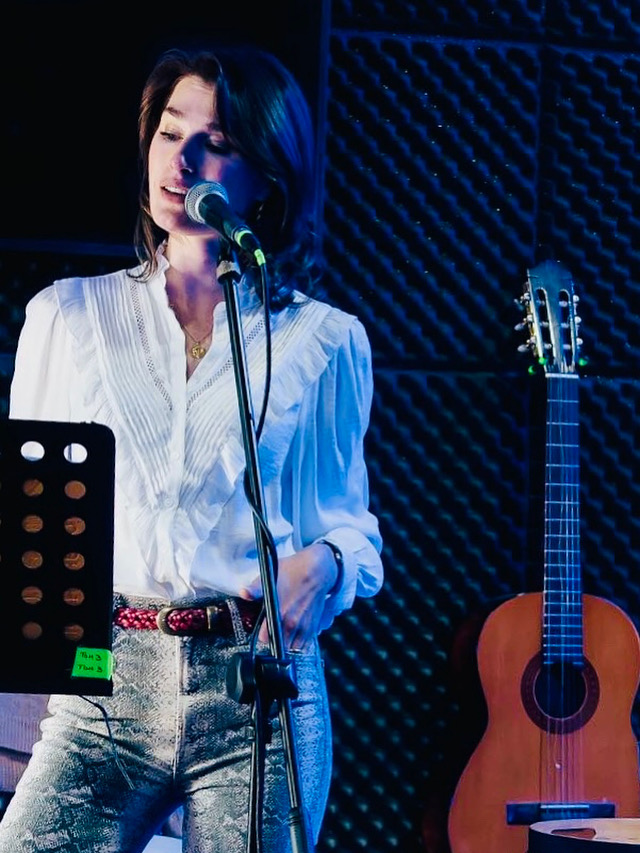 Woman singing into microphone in a recording studio with acoustic foam panels, guitar on stand nearby.