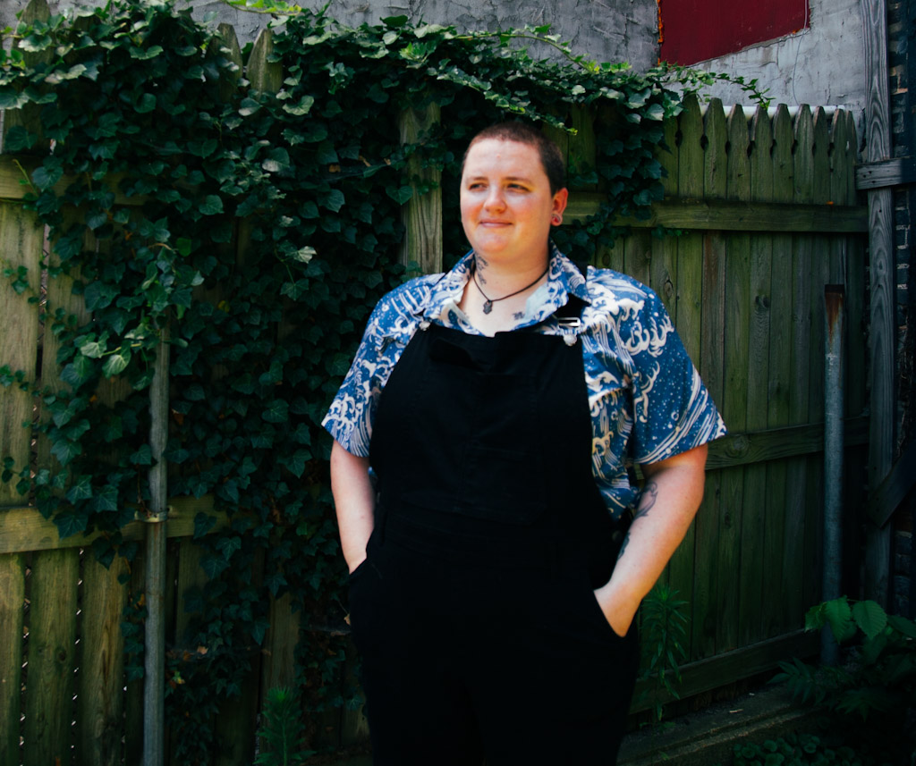 Person standing outdoors in front of a wooden fence and greenery, smiling, wearing a black outfit and patterned shirt, hands in pockets.