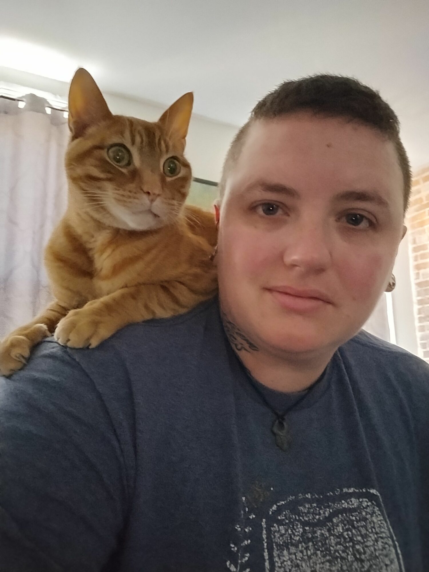 Person with short dark hair holding an orange cat with green eyes, both looking at the camera, indoors with curtains in background.