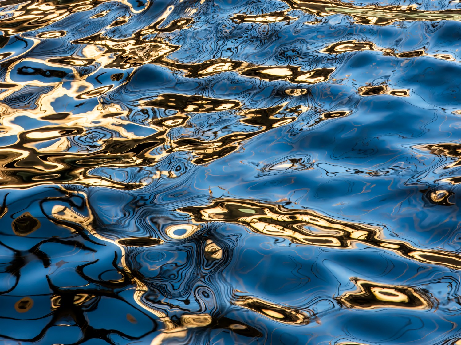 Close-up of water surface with ripples reflecting sunlight, creating a pattern of blue, gold, and black colors.