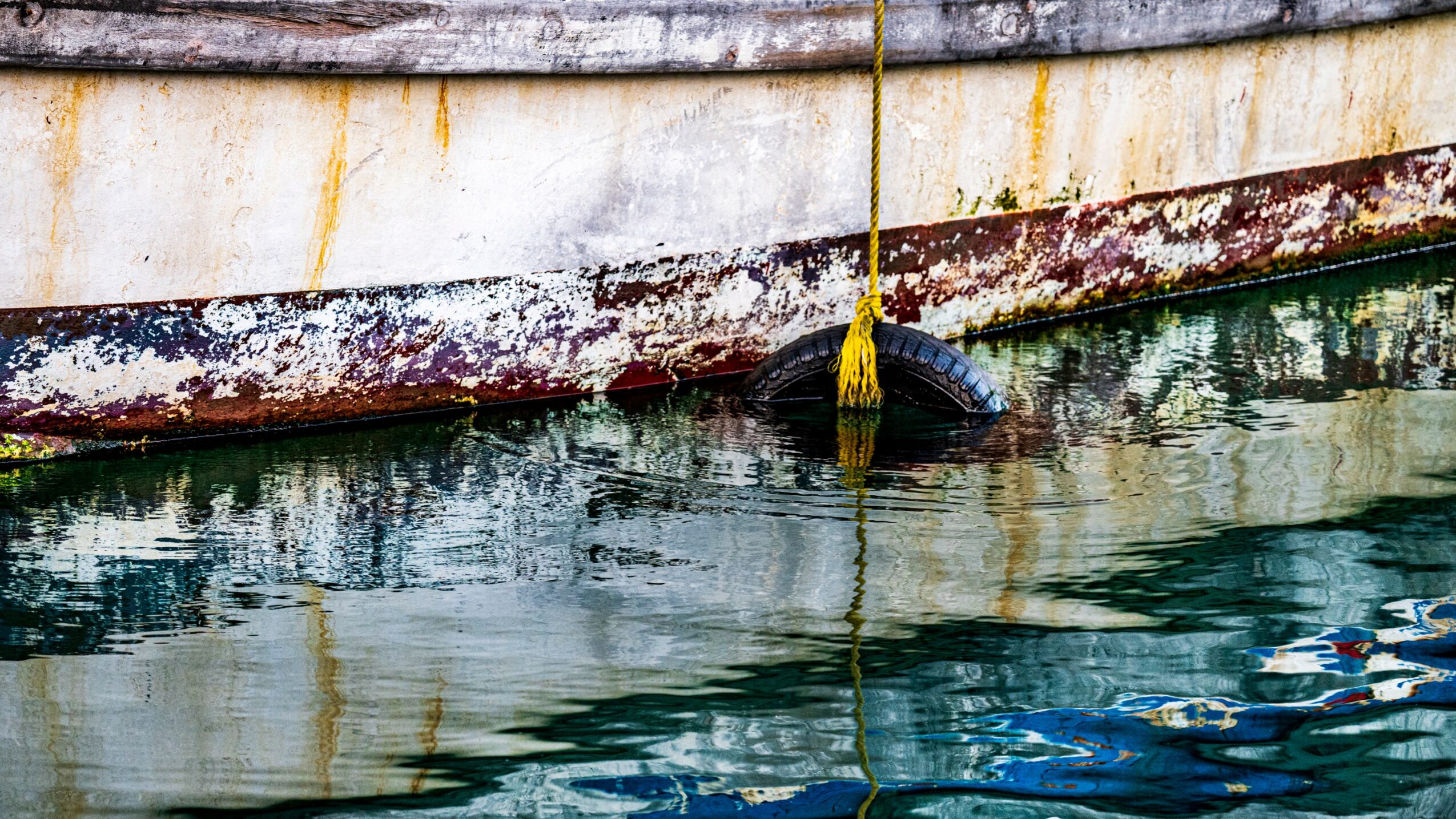 Close-up of a boat's hull with rust and algae, floating on water with a yellow rope hanging down.