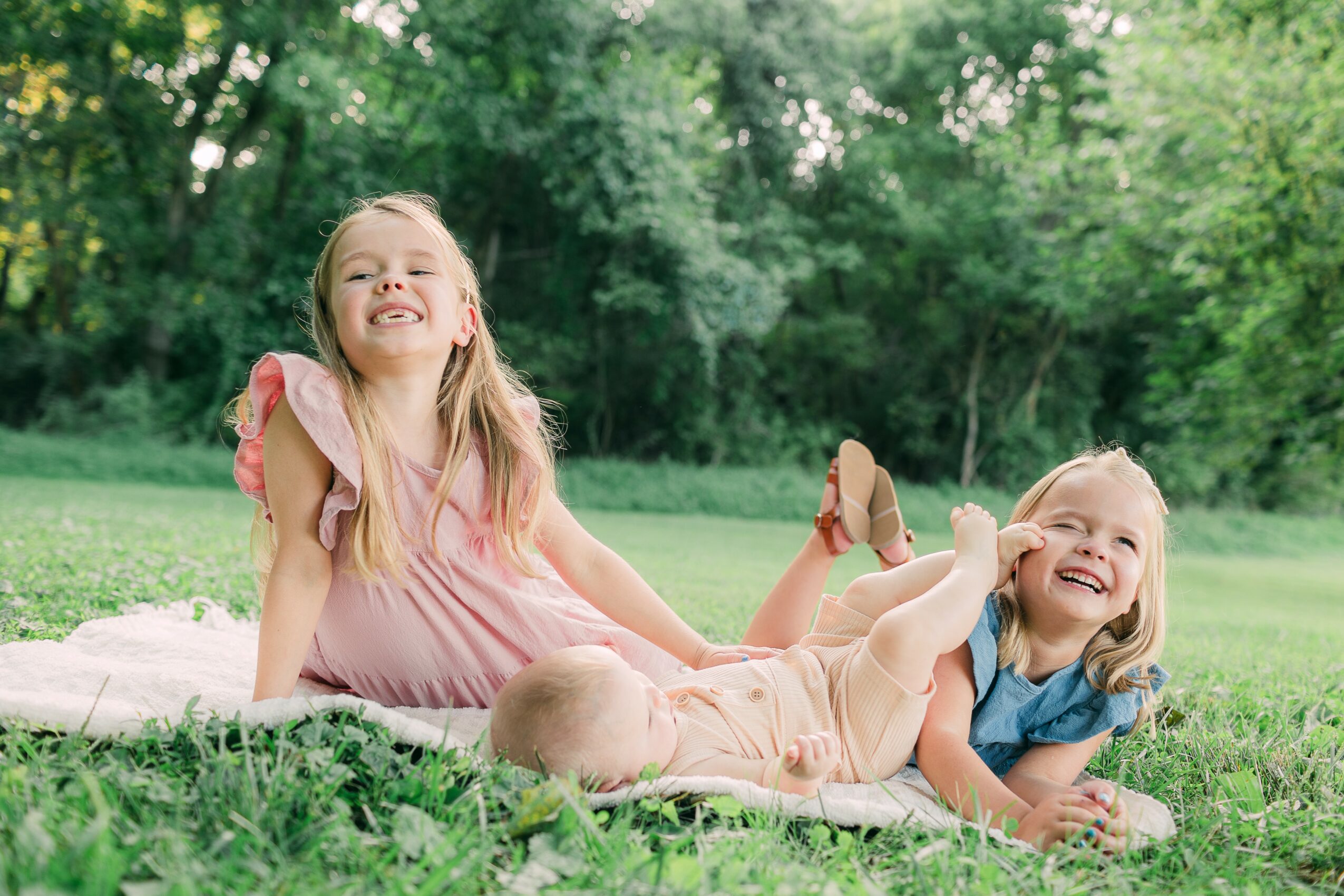 Two young girls lying on grass outdoors, smiling and playing, with trees in the background.