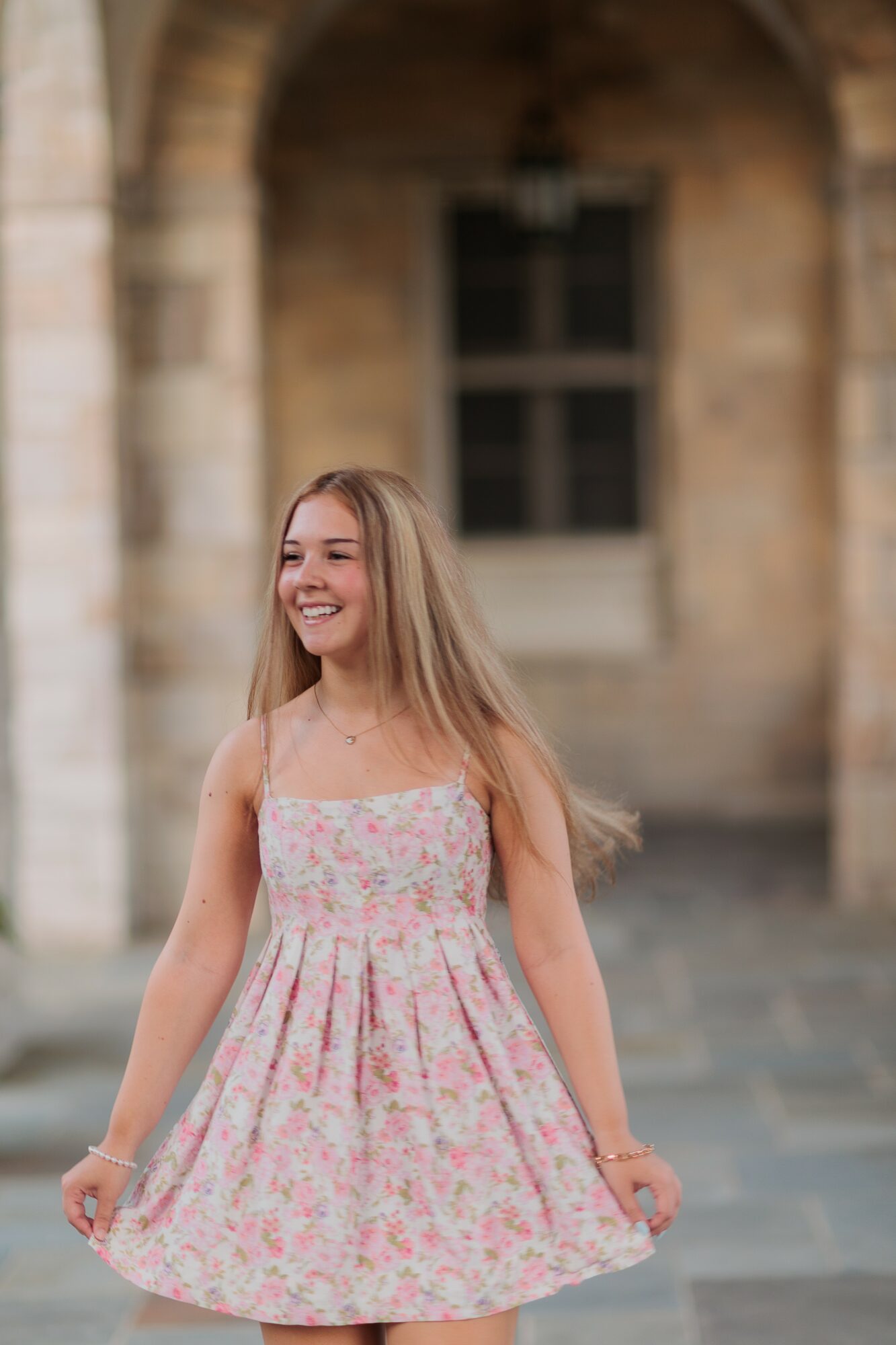 Young woman with long hair smiling, wearing a pink floral dress, standing outdoors in front of a stone building.